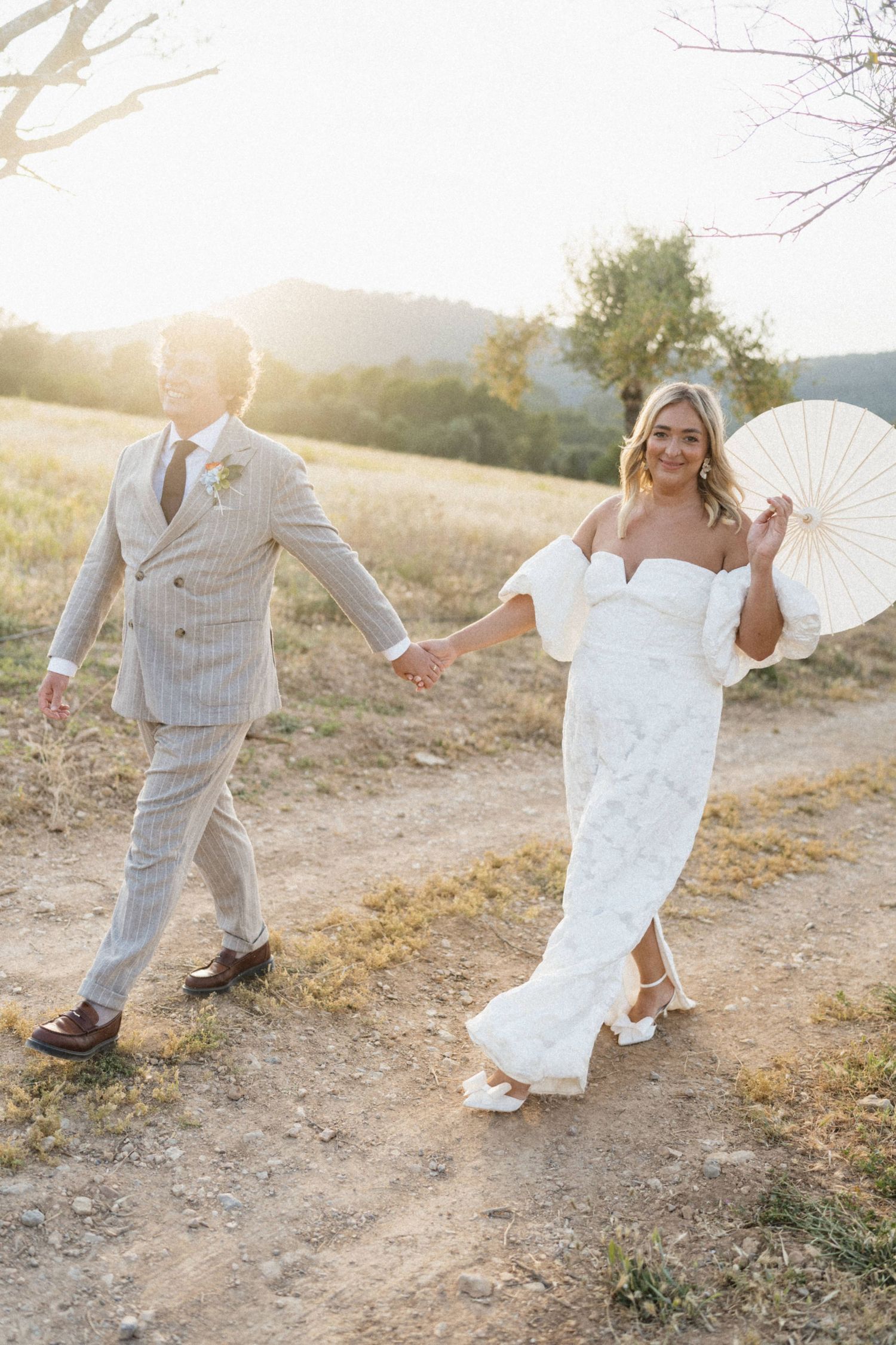 Bride and groom walk hand in hand at golden hour, bride holding white parasol in white gown.