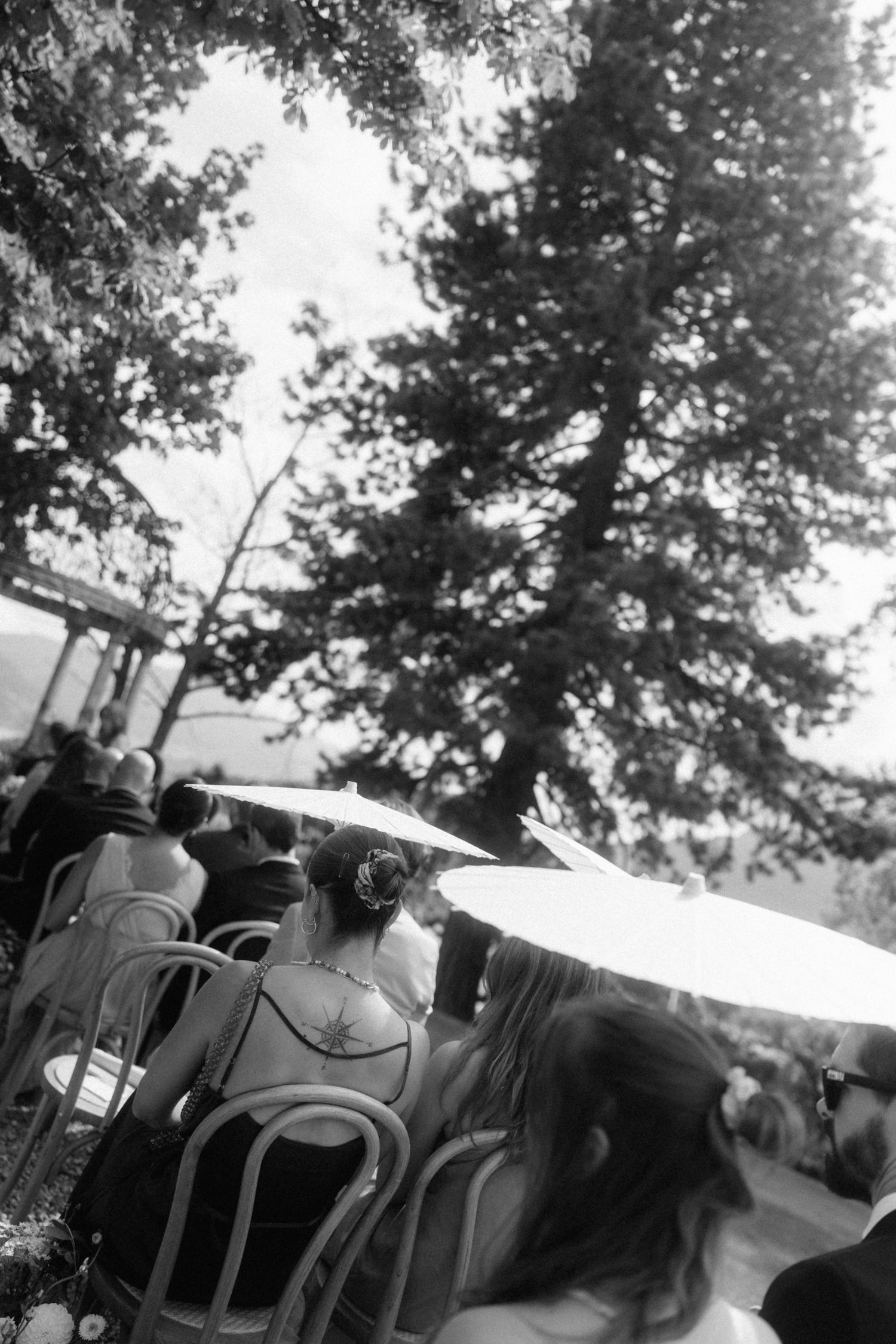 Black and white photo of wedding guests seated outdoors under trees, with a bride holding a parasol visible from behind.