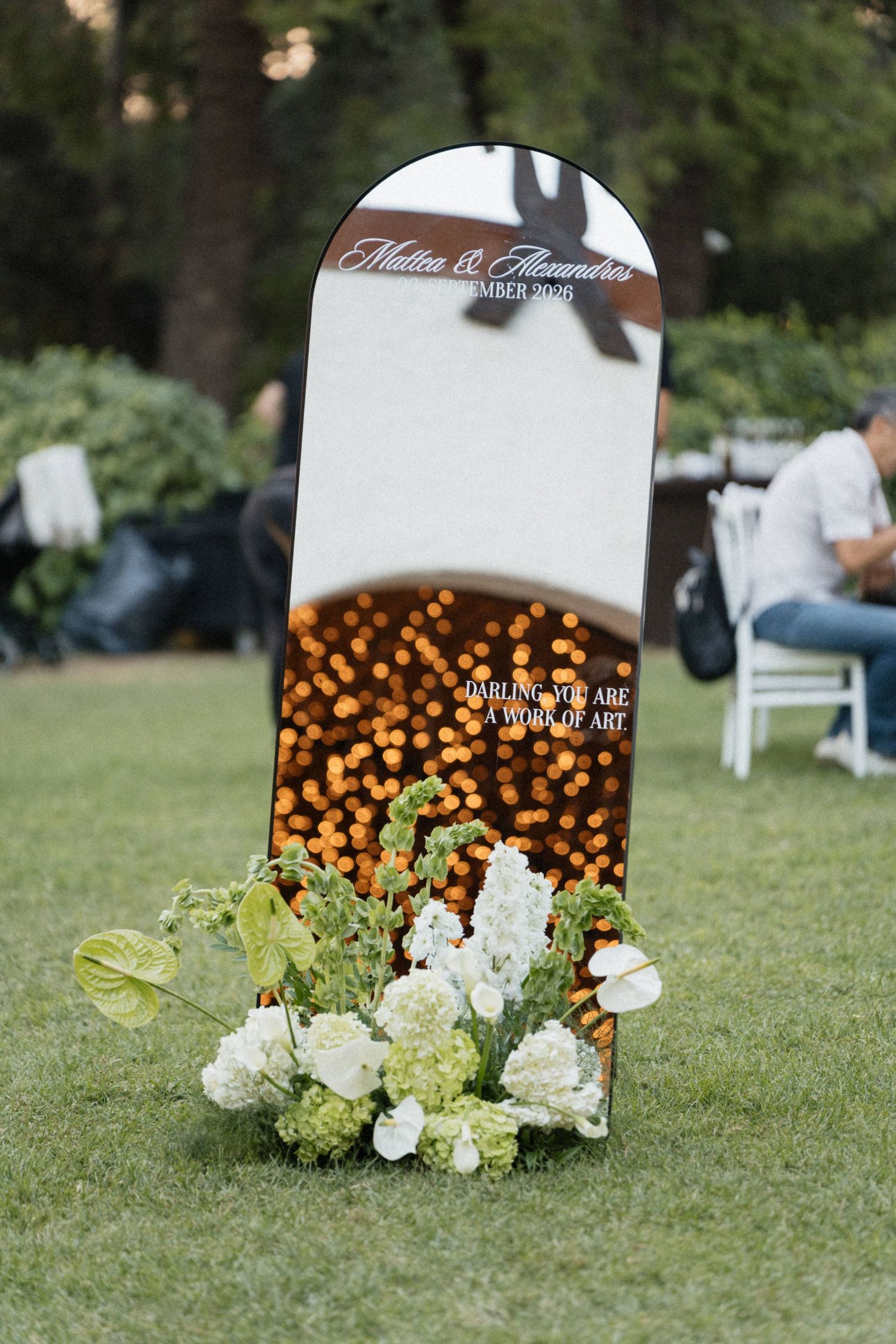 Wedding welcome mirror sign with white and green floral arrangement on grass at outdoor ceremony.