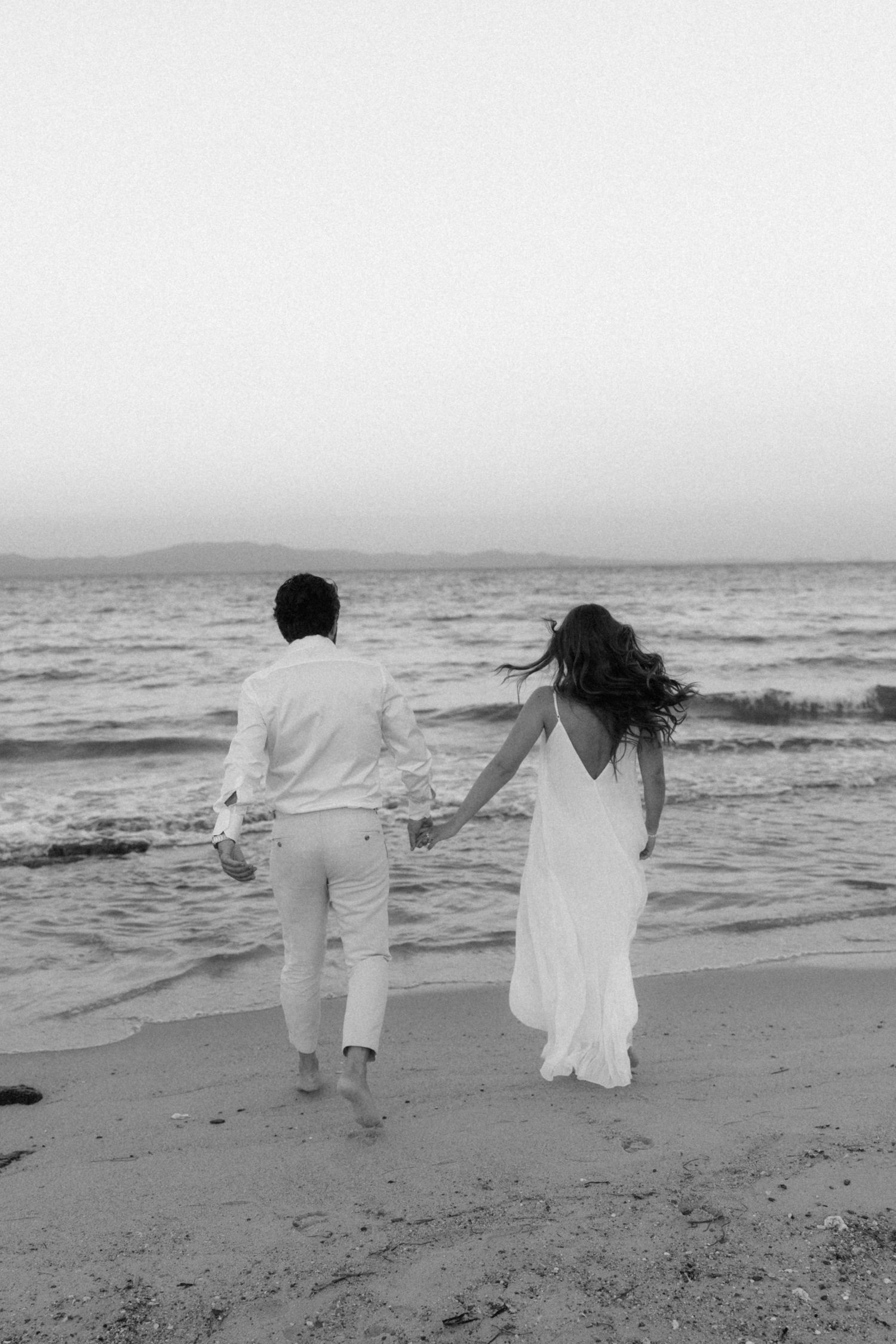Black and white photo of bride and groom holding hands walking toward ocean waves on beach at sunset.