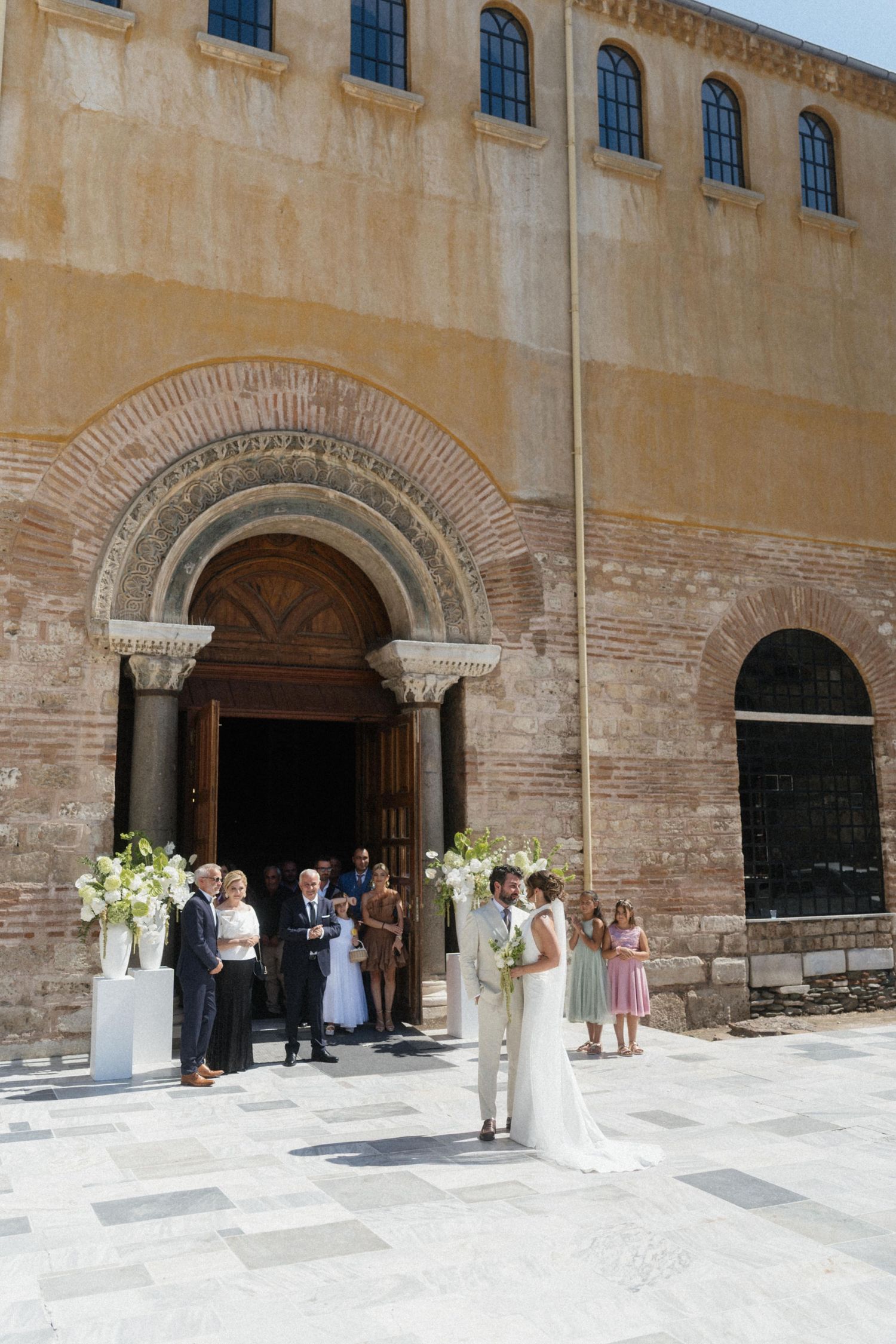 Couple exchanging vows outside a historic stone church adorned with white floral arrangements.