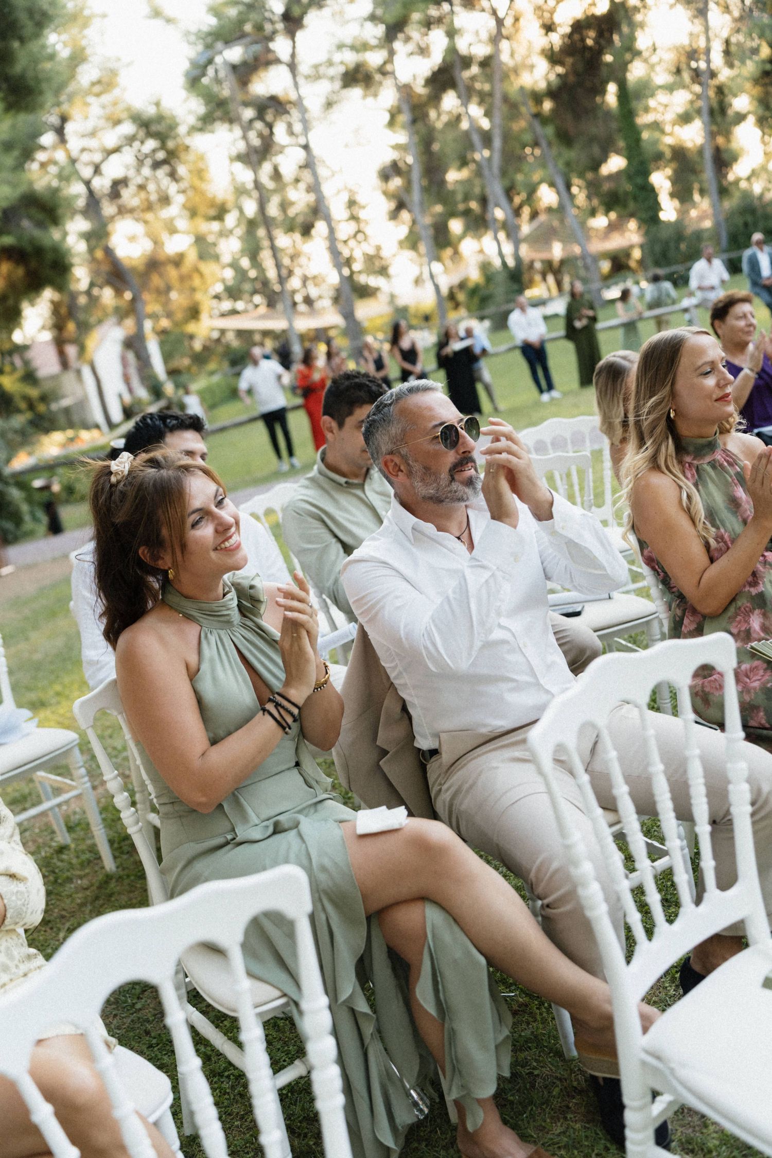 Guests seated on white chairs at an outdoor wedding ceremony in a lush garden, smiling and enjoying the celebration.