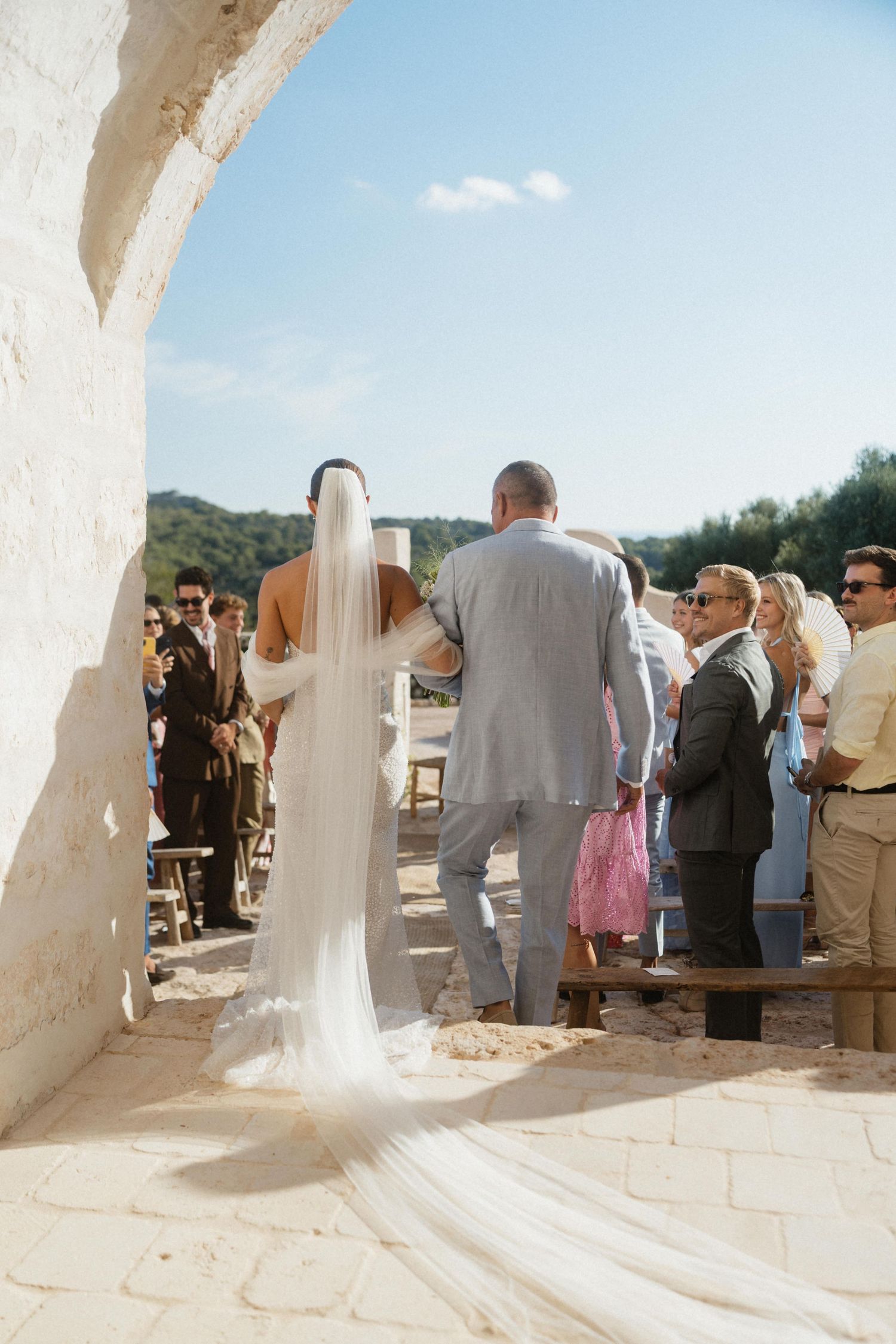 Bride in a long flowing veil walks down the aisle on her father's arm toward guests at a Mediterranean stone ceremony.