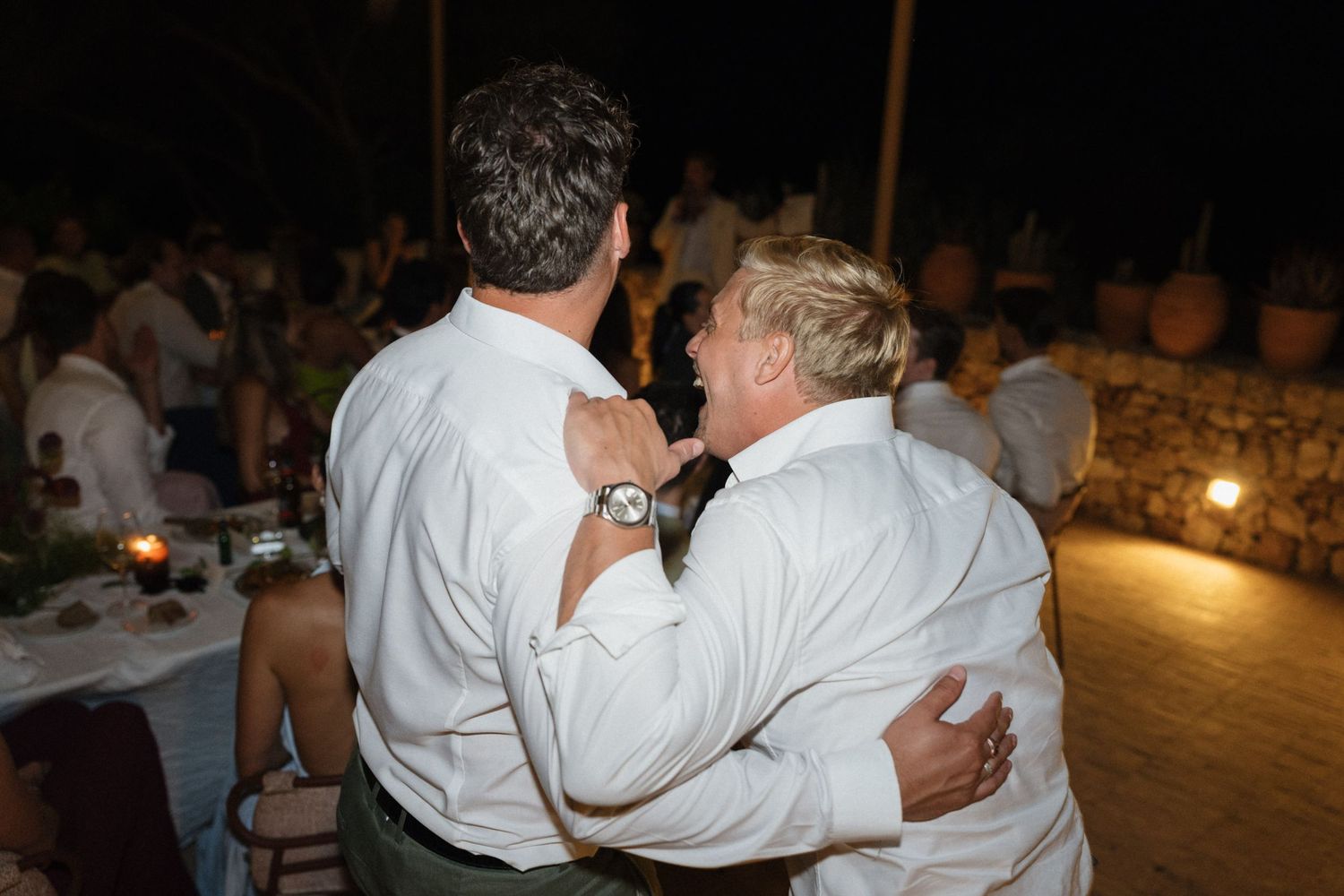 Two men in white shirts share a joyful embrace on the dance floor at a nighttime wedding reception with guests behind them.