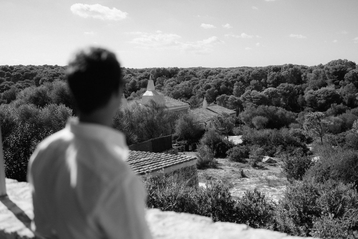 Black and white photo of a man seen from behind gazing over a dense tree-covered landscape with rooftops and a small tower in the distance.
