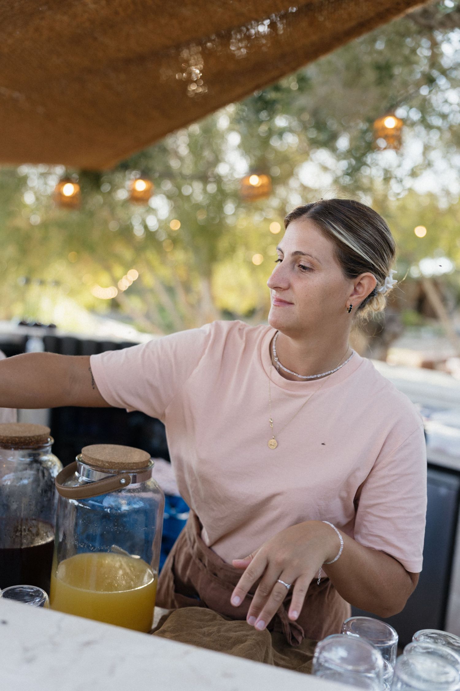 Woman in a pink t-shirt stands behind an outdoor bar with jars of fresh juice and glasses under string lights.