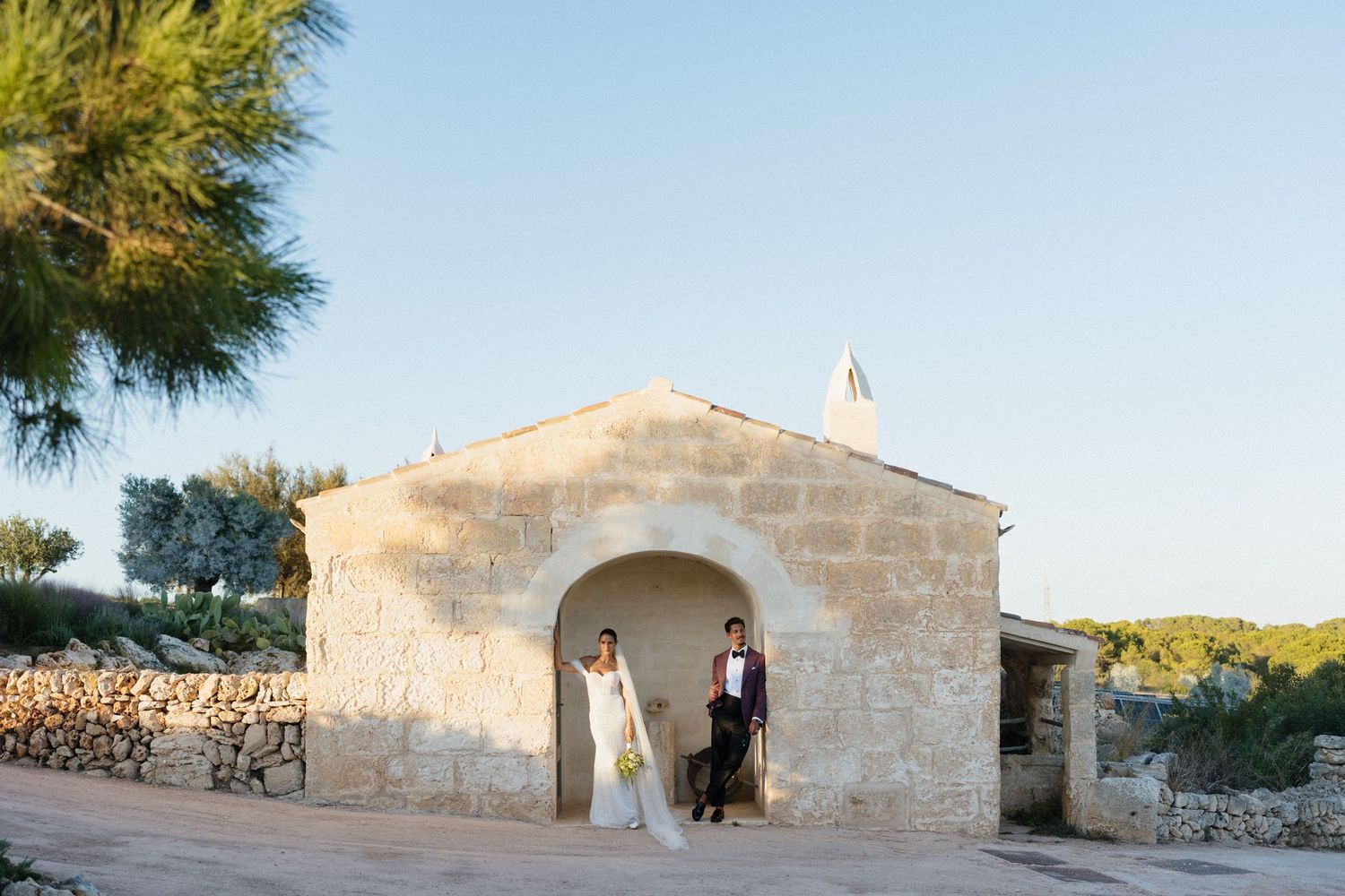Bride in white gown and groom in dark suit pose in the arched doorway of a rustic stone chapel with pine trees and a blue sky.