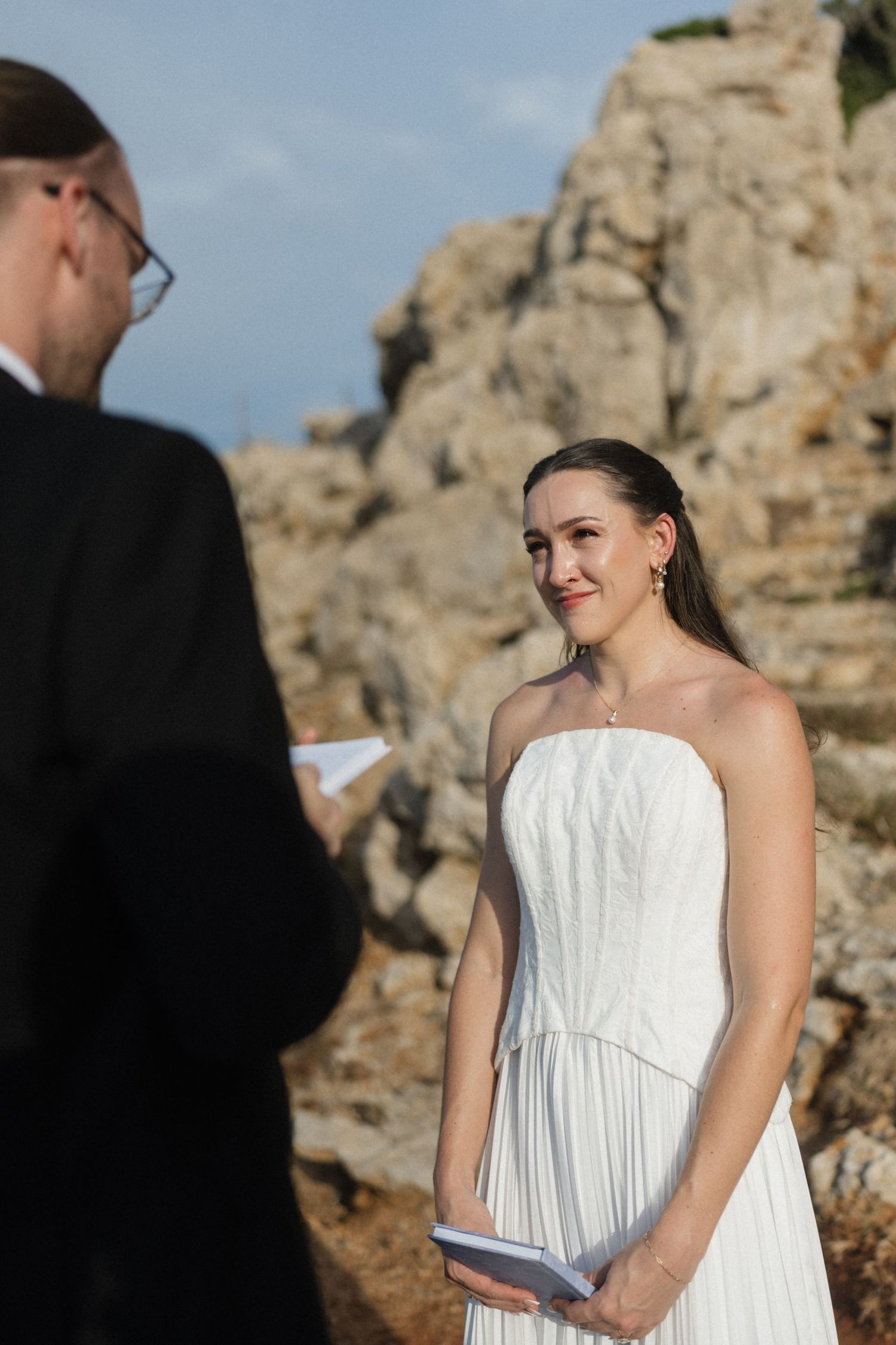 Smiling bride in a white pleated strapless gown holds a small book while facing her partner during an outdoor ceremony by rocky cliffs.