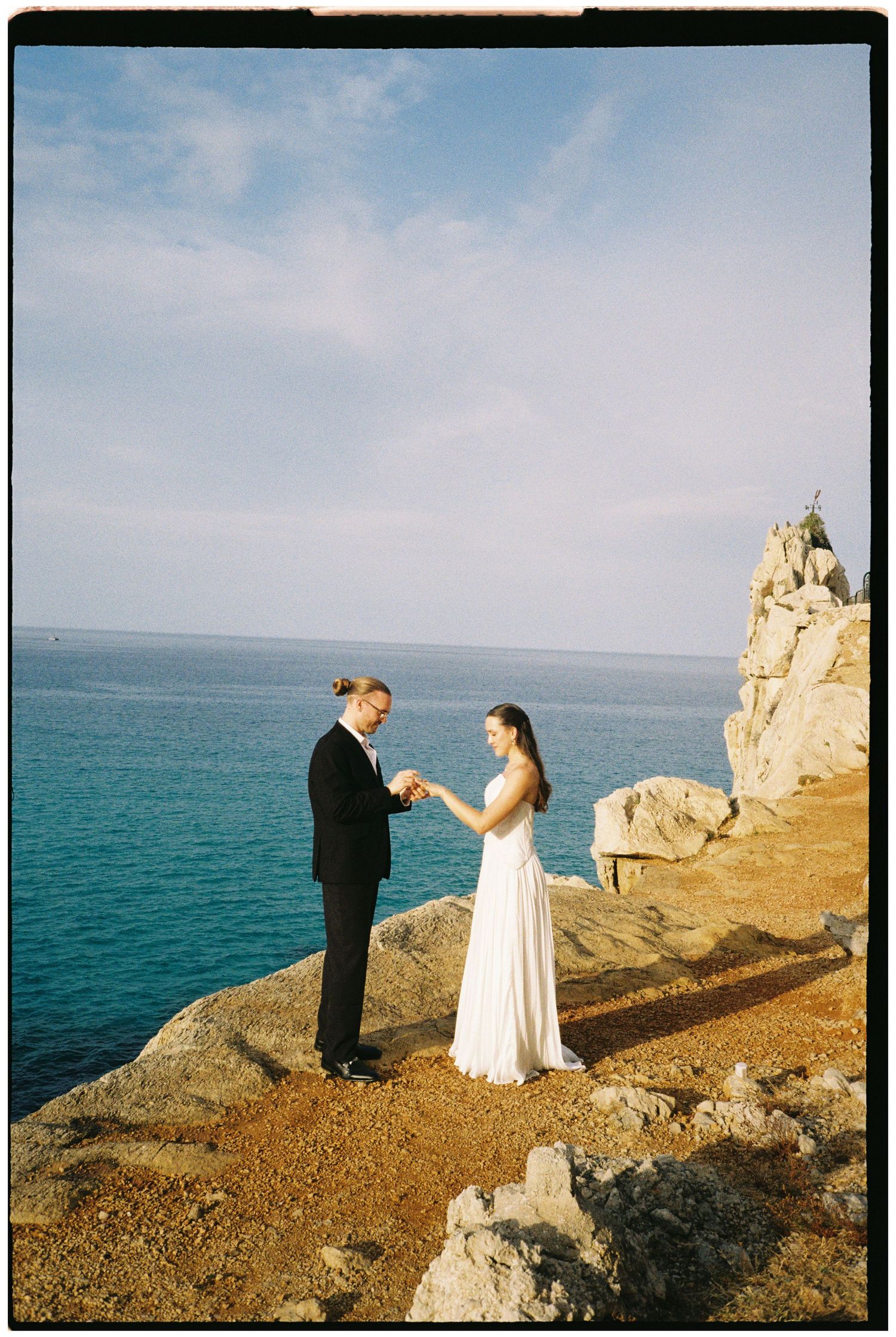 Bride in a white gown and groom in a dark suit exchange rings on a rocky seaside cliff overlooking the turquoise ocean at sunset.