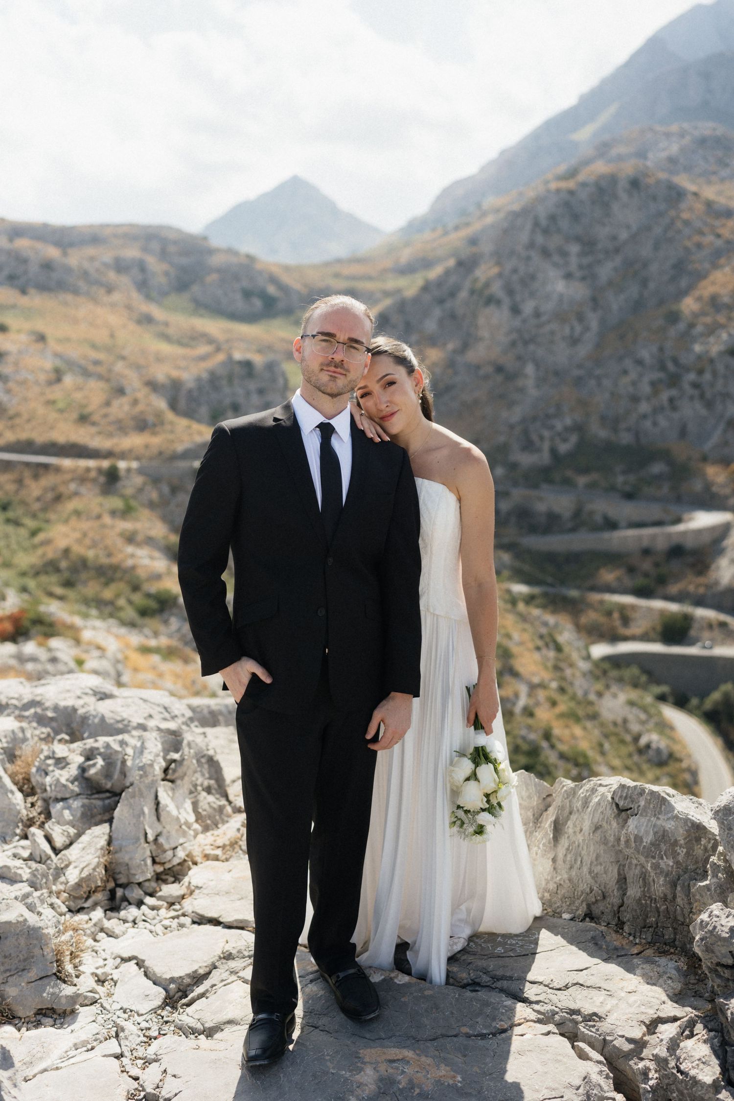 Bride in a white strapless gown holding a bouquet leans on her groom in a black suit on rocky cliffs above a winding mountain road.