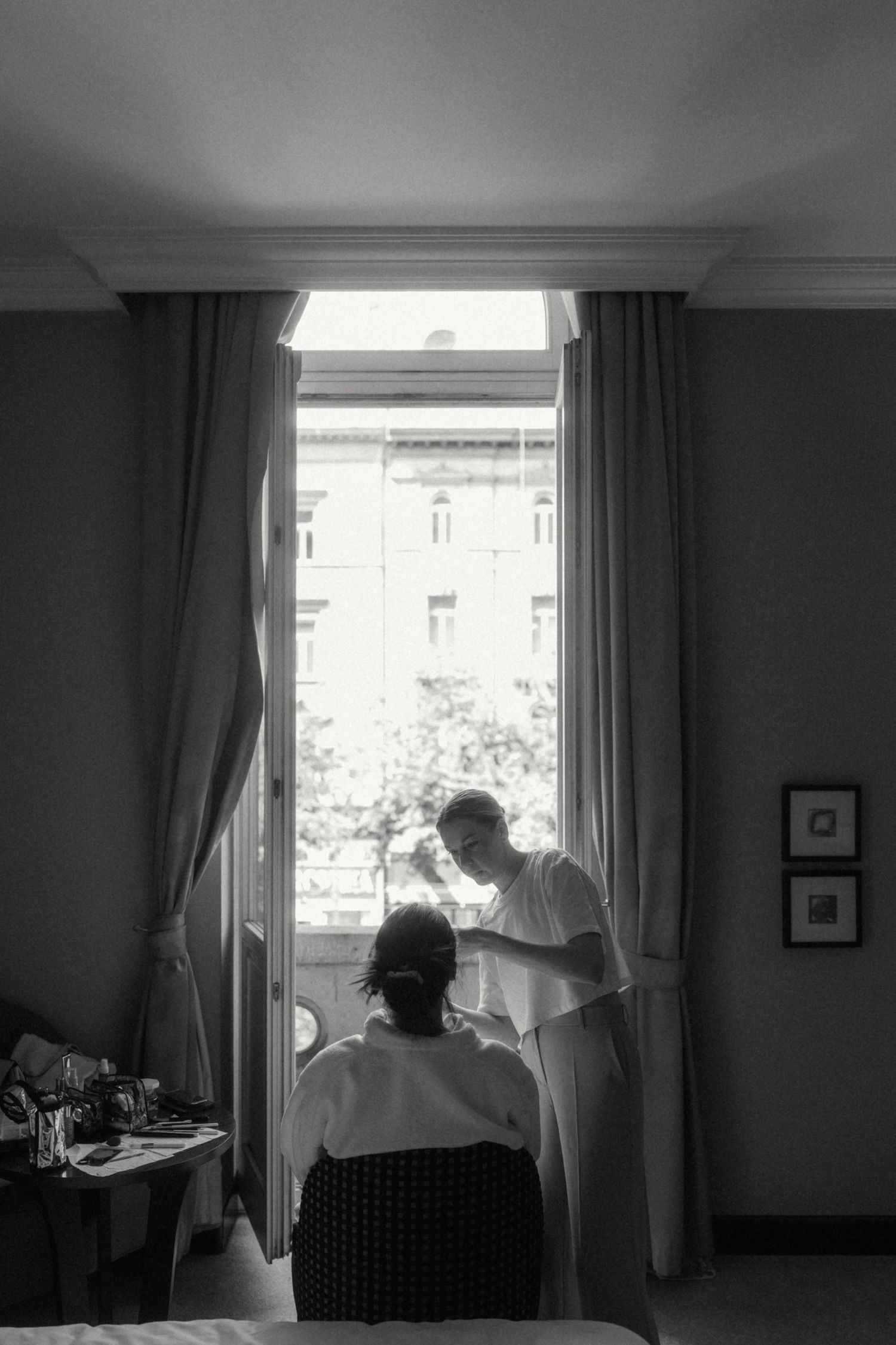 Black and white photo of bride getting ready near window with natural light streaming in.