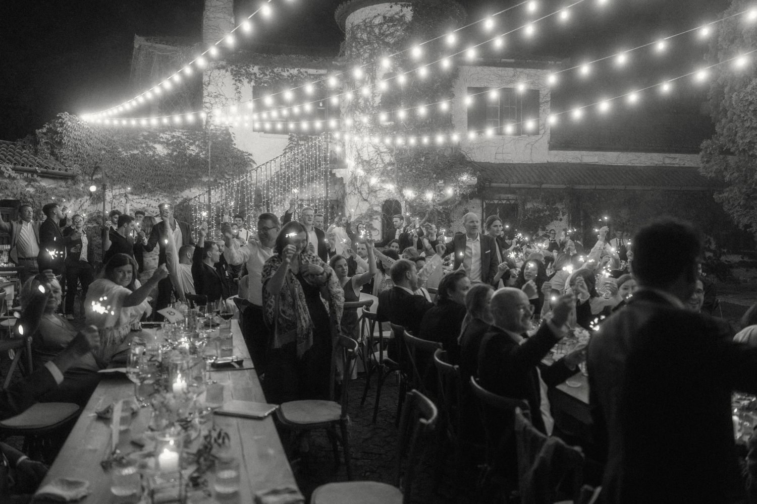 Black and white photo of wedding guests raising glasses and phones under string lights at a night reception.