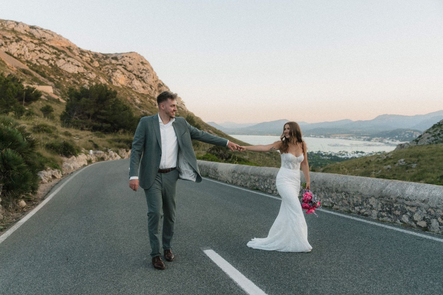Bride and groom walk hand in hand down a scenic mountain road at sunset, bride in white gown holding pink flowers.