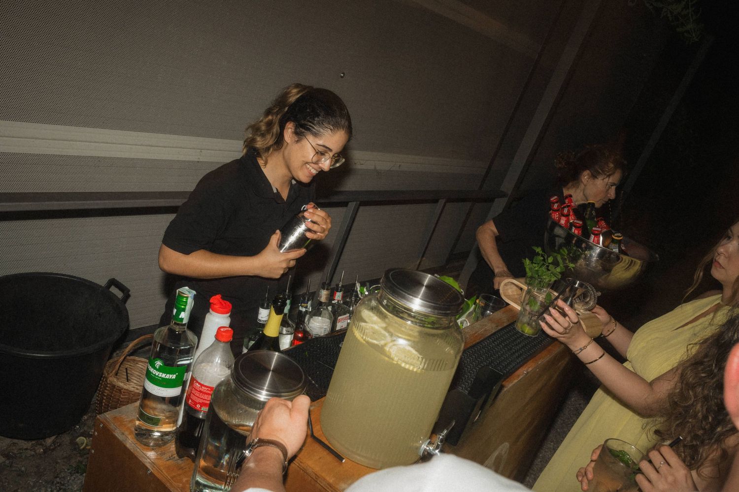 Bartender in black outfit preparing drinks at an outdoor wedding reception bar at night.