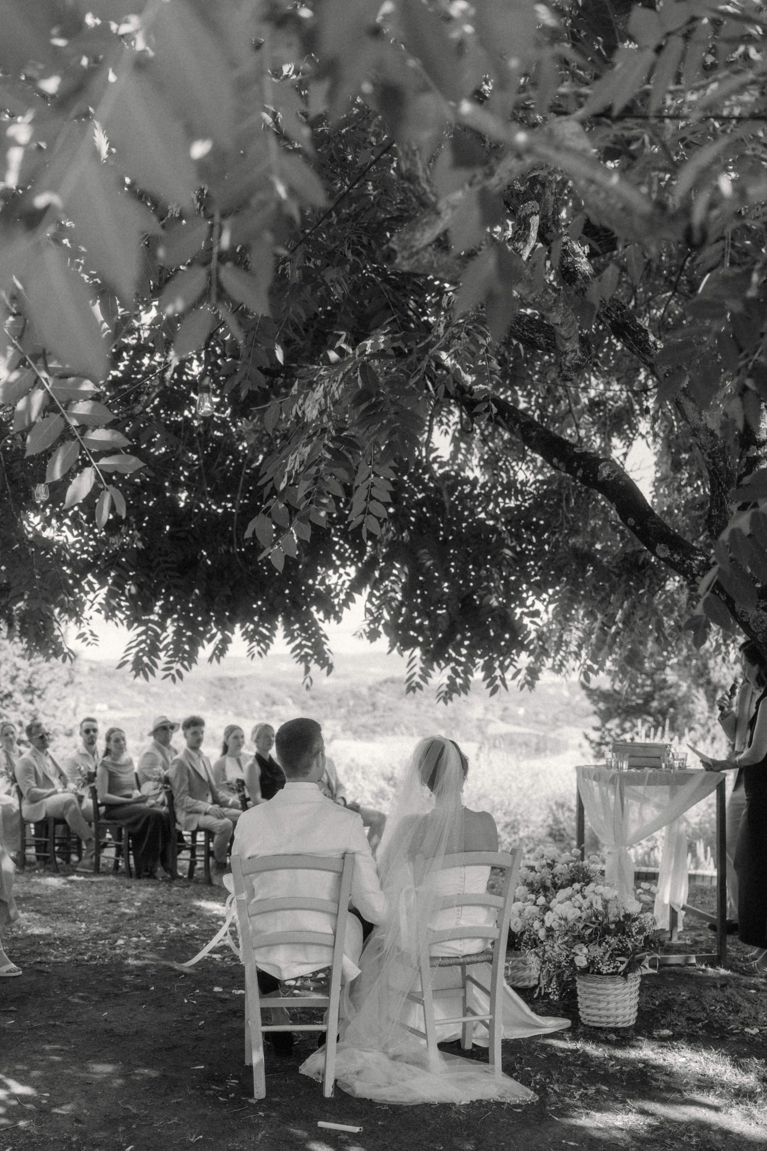 Black and white photo of bride and groom seated at outdoor wedding ceremony under lush tree canopy.