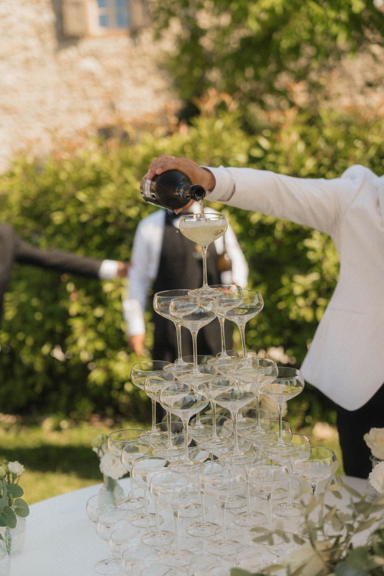 Waiter pouring champagne into crystal coupe glass tower at outdoor wedding reception.