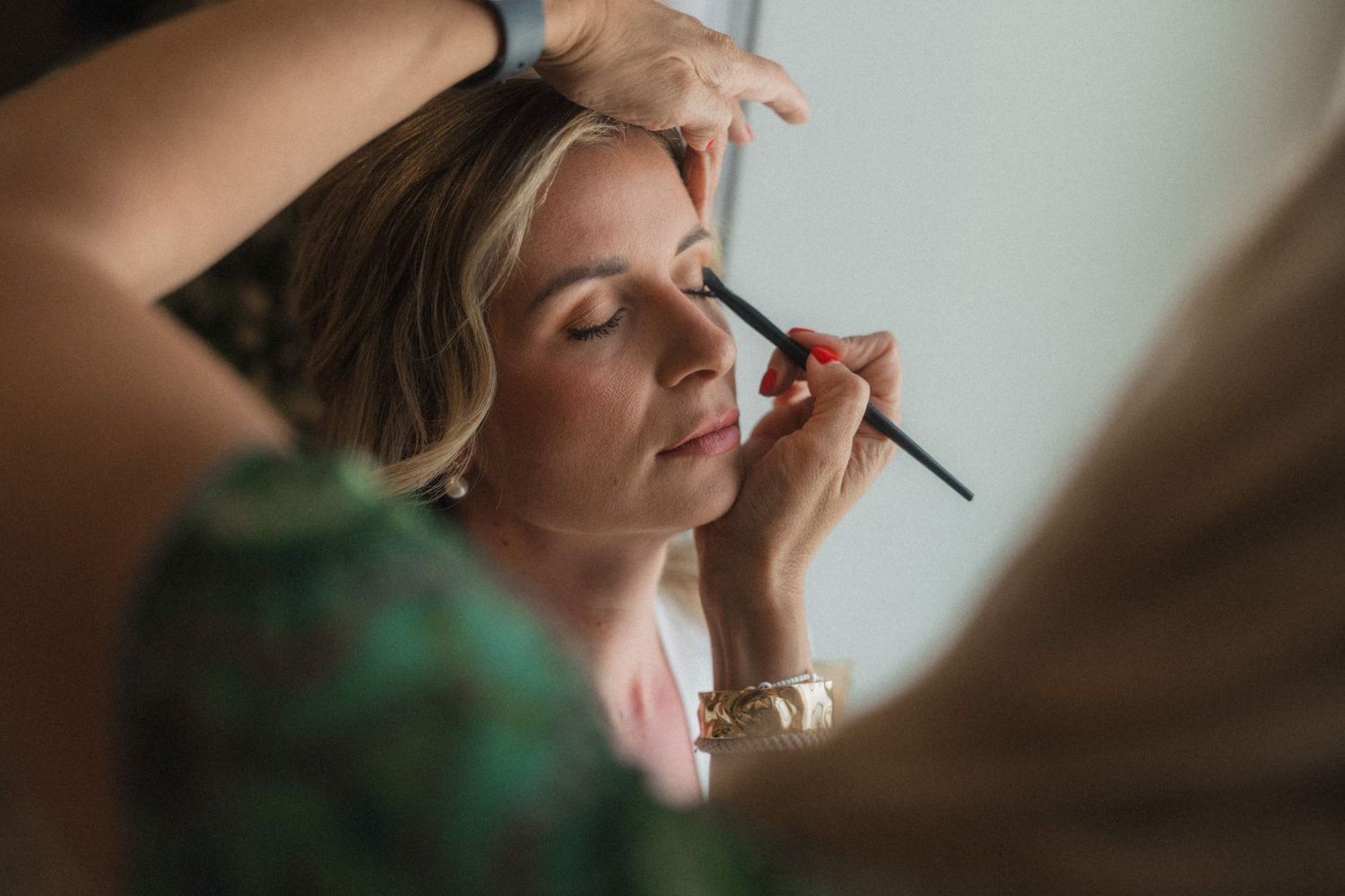 Professional makeup artist applying eye makeup to a blonde woman with a brush.