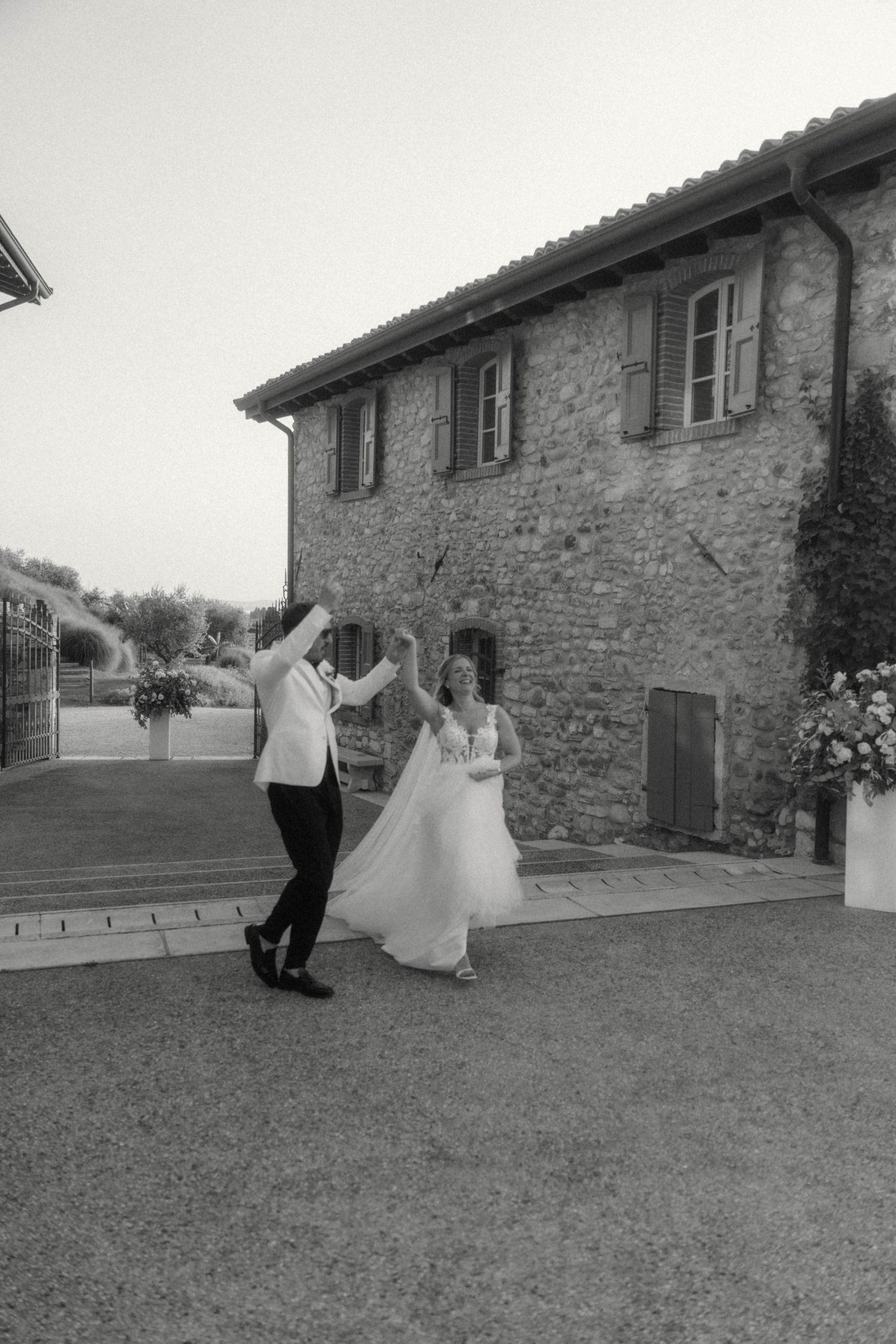 Black and white photo of bride and groom dancing joyfully outside rustic stone villa.