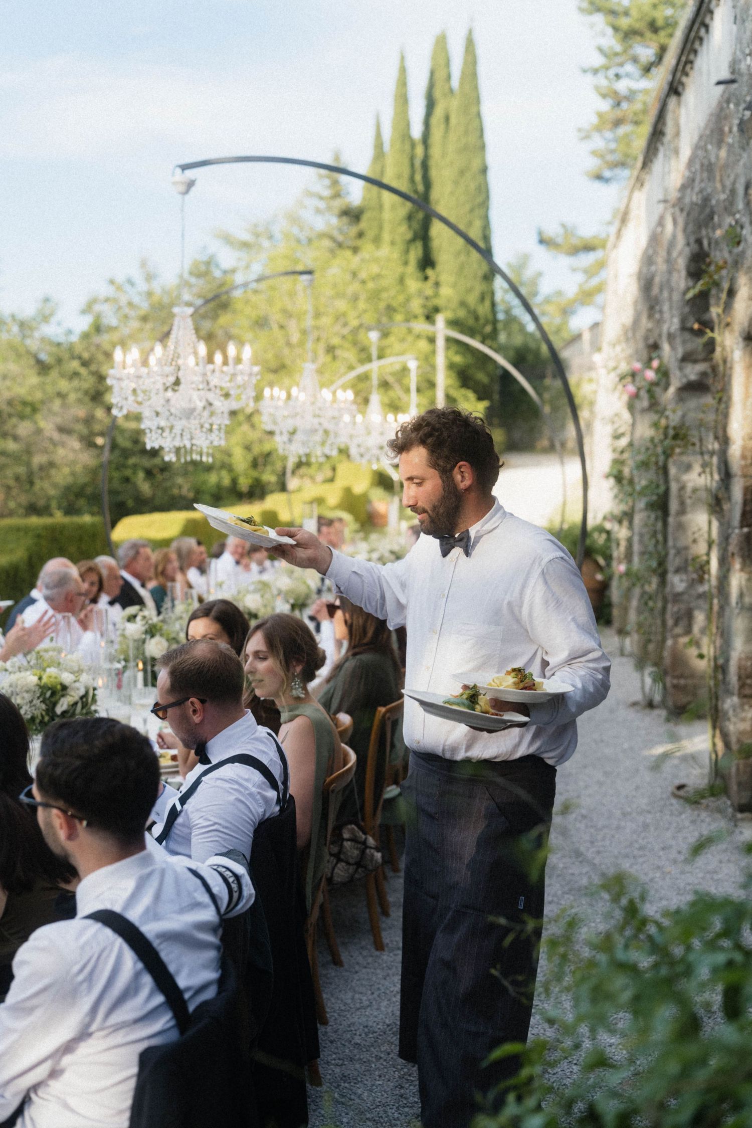 Waiter serving plates at an elegant outdoor wedding reception with crystal chandeliers and long dining tables.