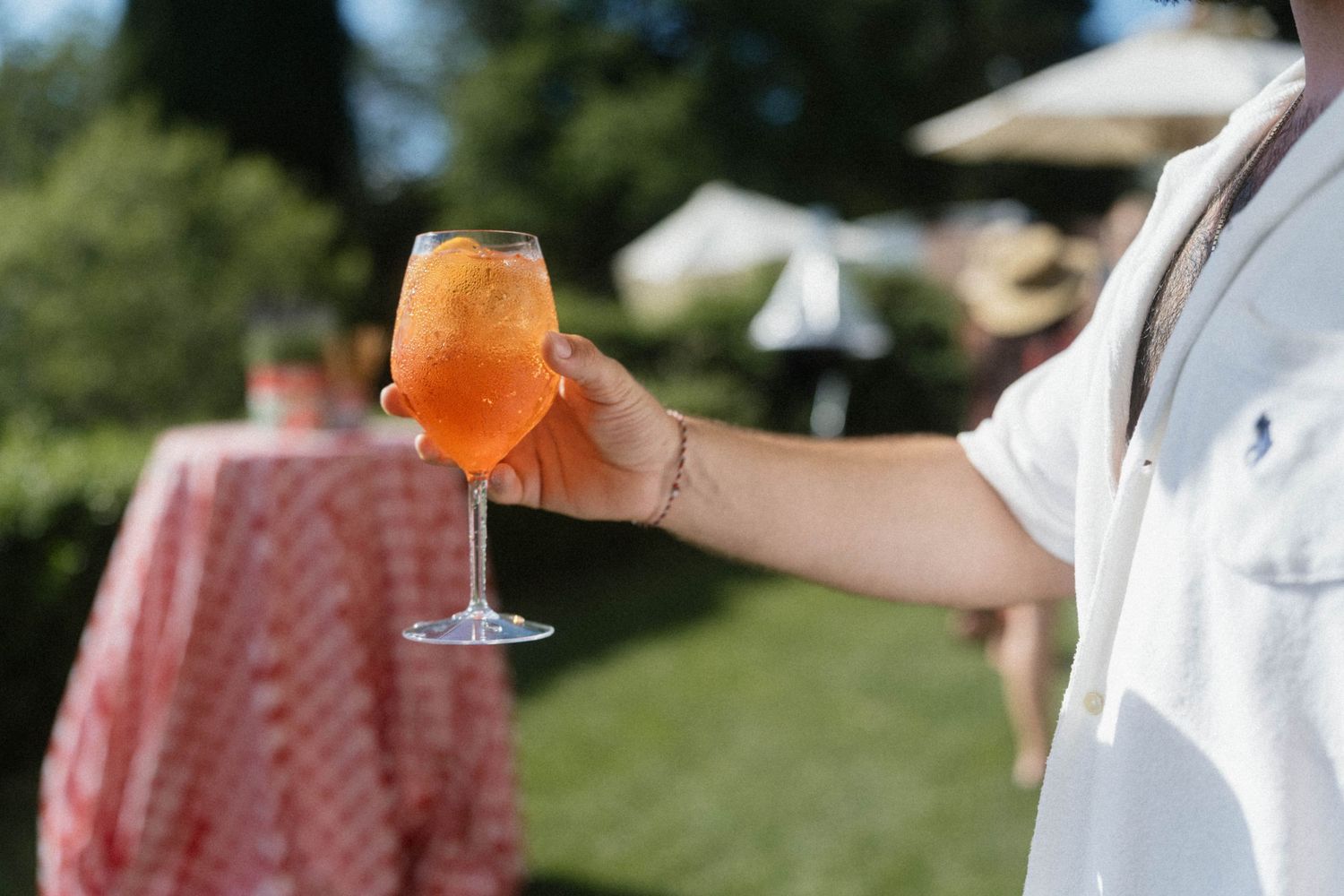 Hand holding an Aperol Spritz cocktail at an outdoor garden wedding reception.