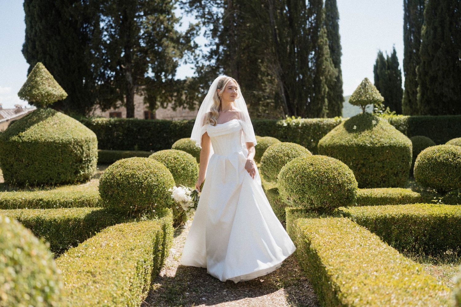 Elegant bride in white ball gown and veil poses in formal Italian garden with sculpted hedges.