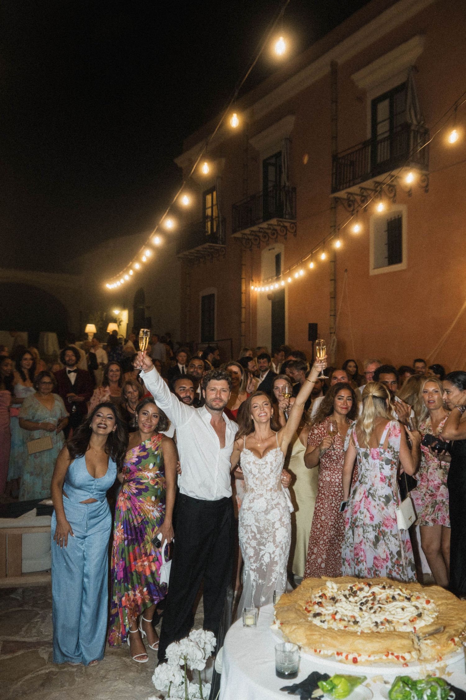 Bride and groom toast with champagne surrounded by wedding guests under string lights at night outdoor reception.