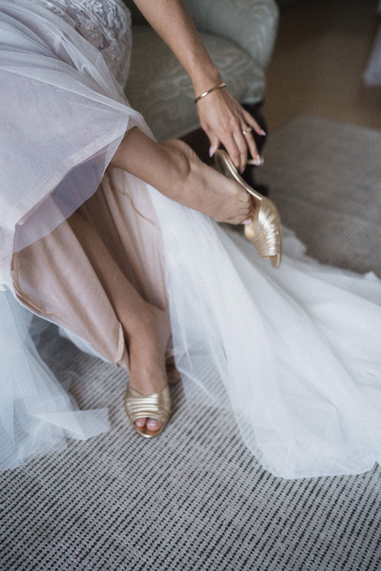 Bride sitting and putting on gold strappy heels beneath her white tulle wedding gown.