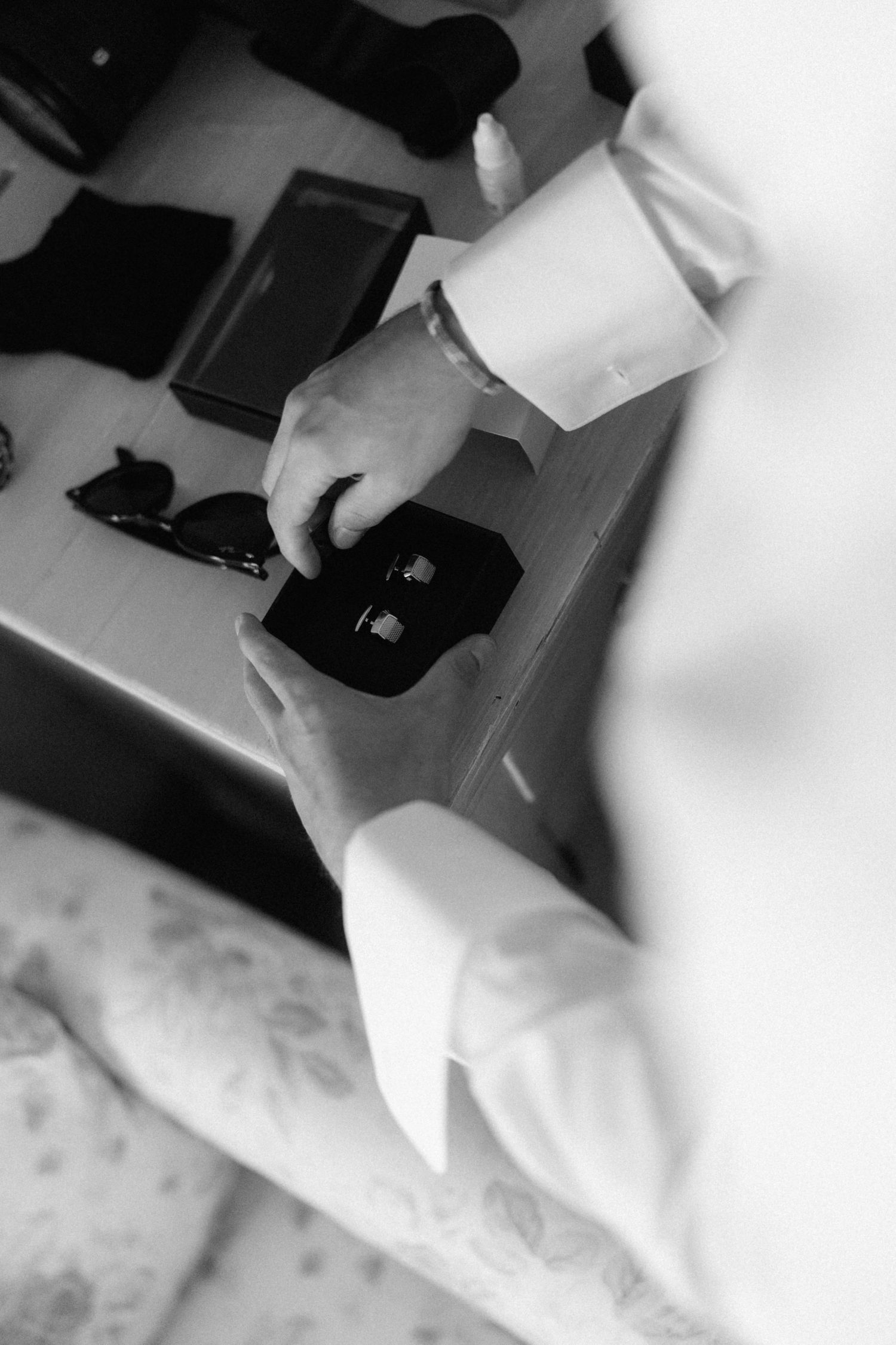Black and white close-up of groom selecting cufflinks from a box on a dresser.