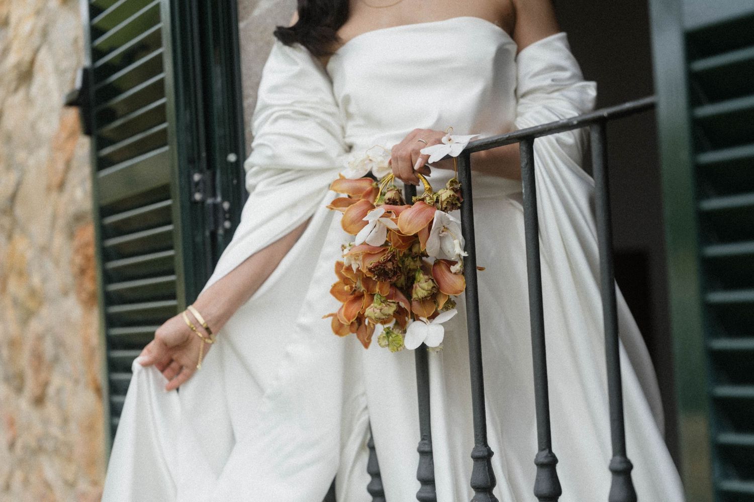 Bride in off-shoulder white wedding gown holding autumn floral bouquet on stone steps.