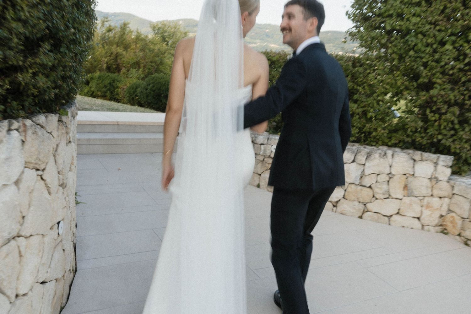 A bride in a white gown and veil walks with a groom in a black suit on an outdoor stone terrace.