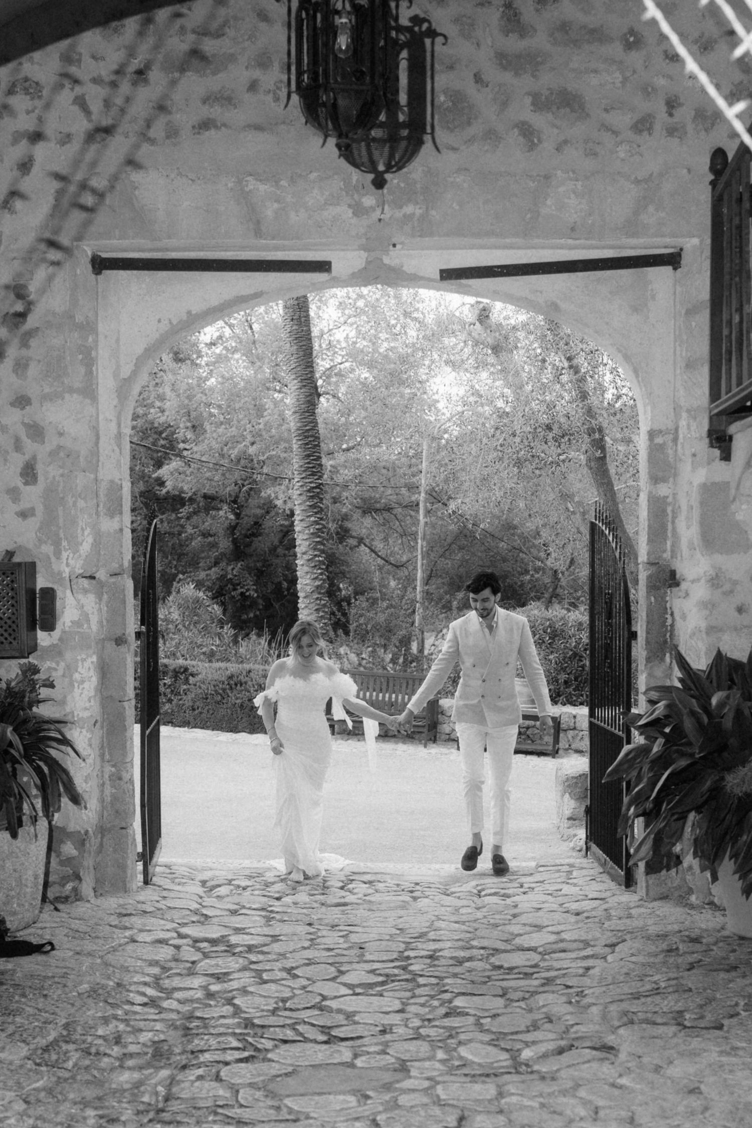 Black and white photo of bride and groom walking hand in hand through stone archway.