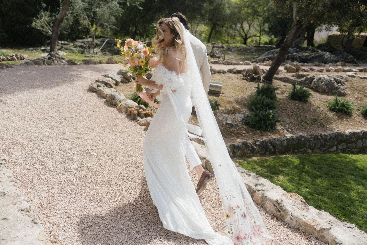 A bride in a flowing white gown with a floral veil walks through a scenic outdoor garden on her wedding day.
