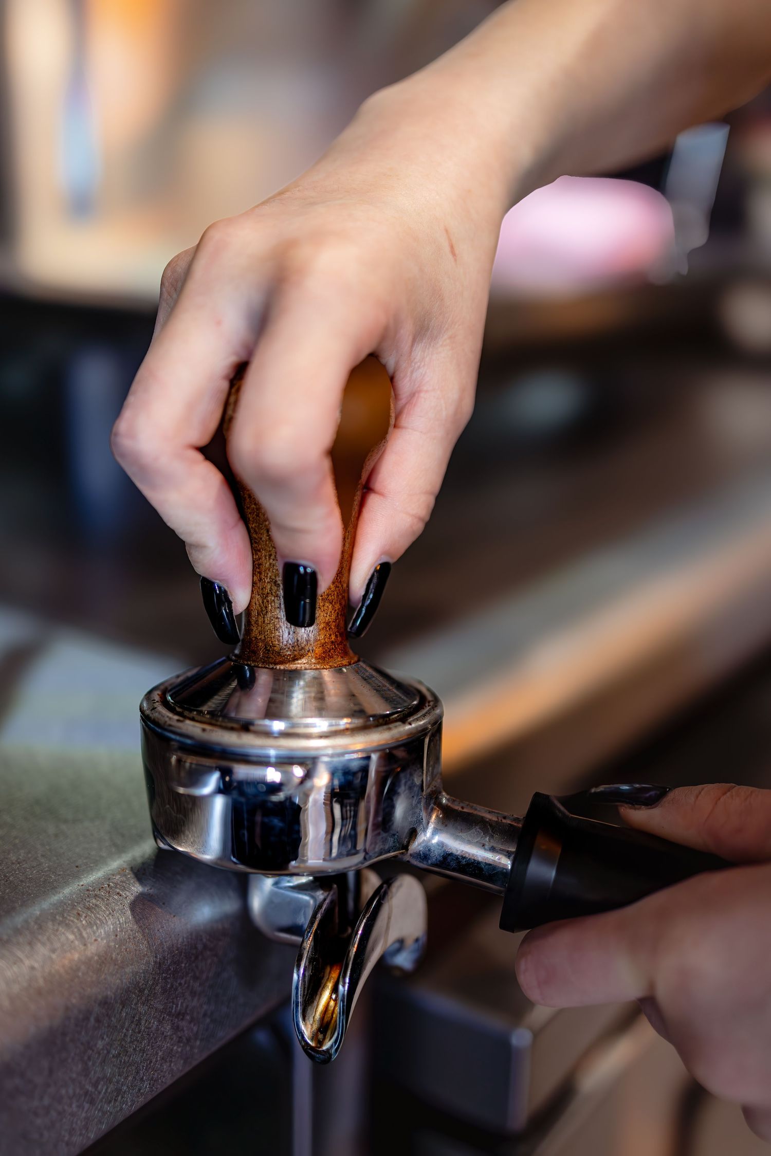 Barista with black nail polish tamping coffee grounds into portafilter at espresso machine.