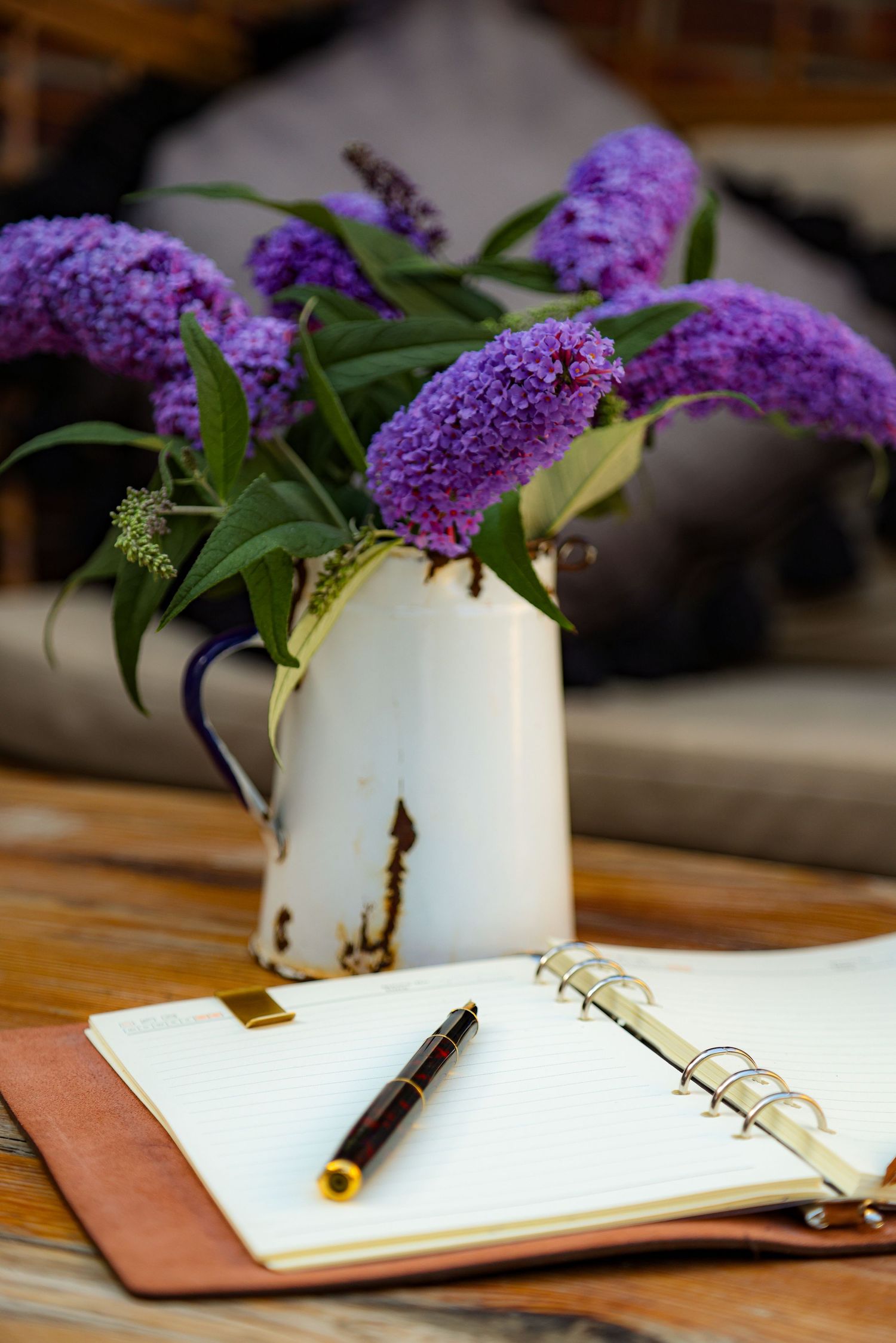 Open notebook with a pen beside a rustic vase of purple butterfly bush flowers on a wooden table.