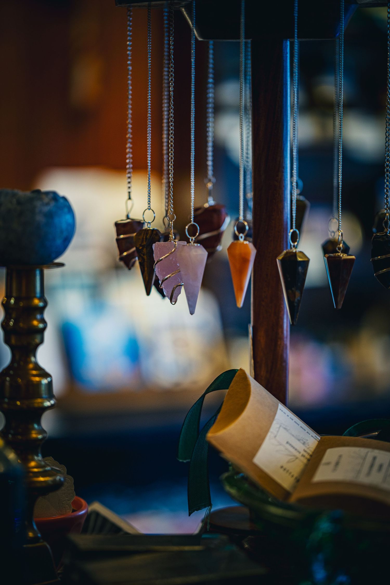 Crystal pendulum necklaces hanging from wooden display with open books and candlestick below.