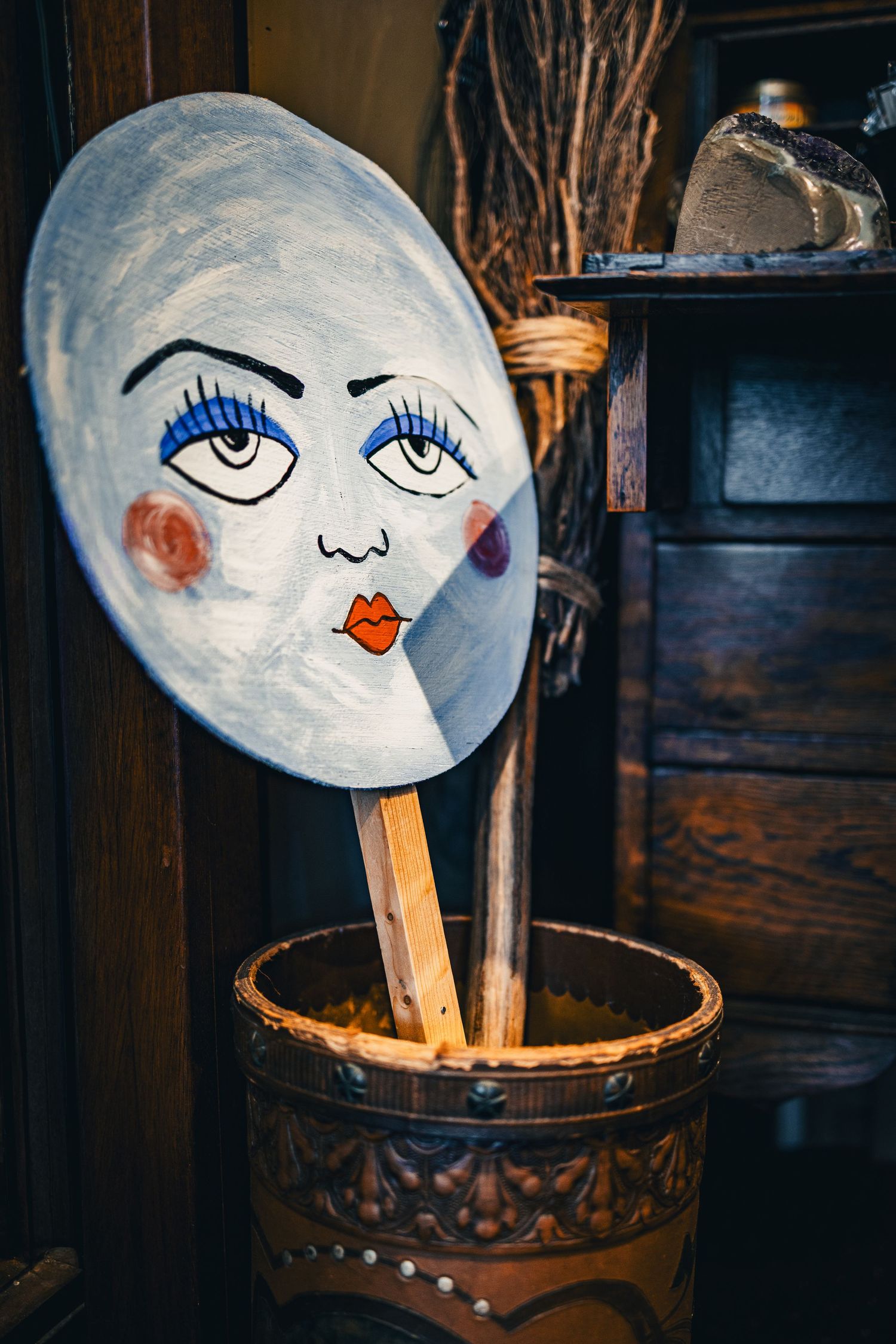 Hand-painted white oval face mask on wooden stick displayed in decorative carved wooden bucket indoors.