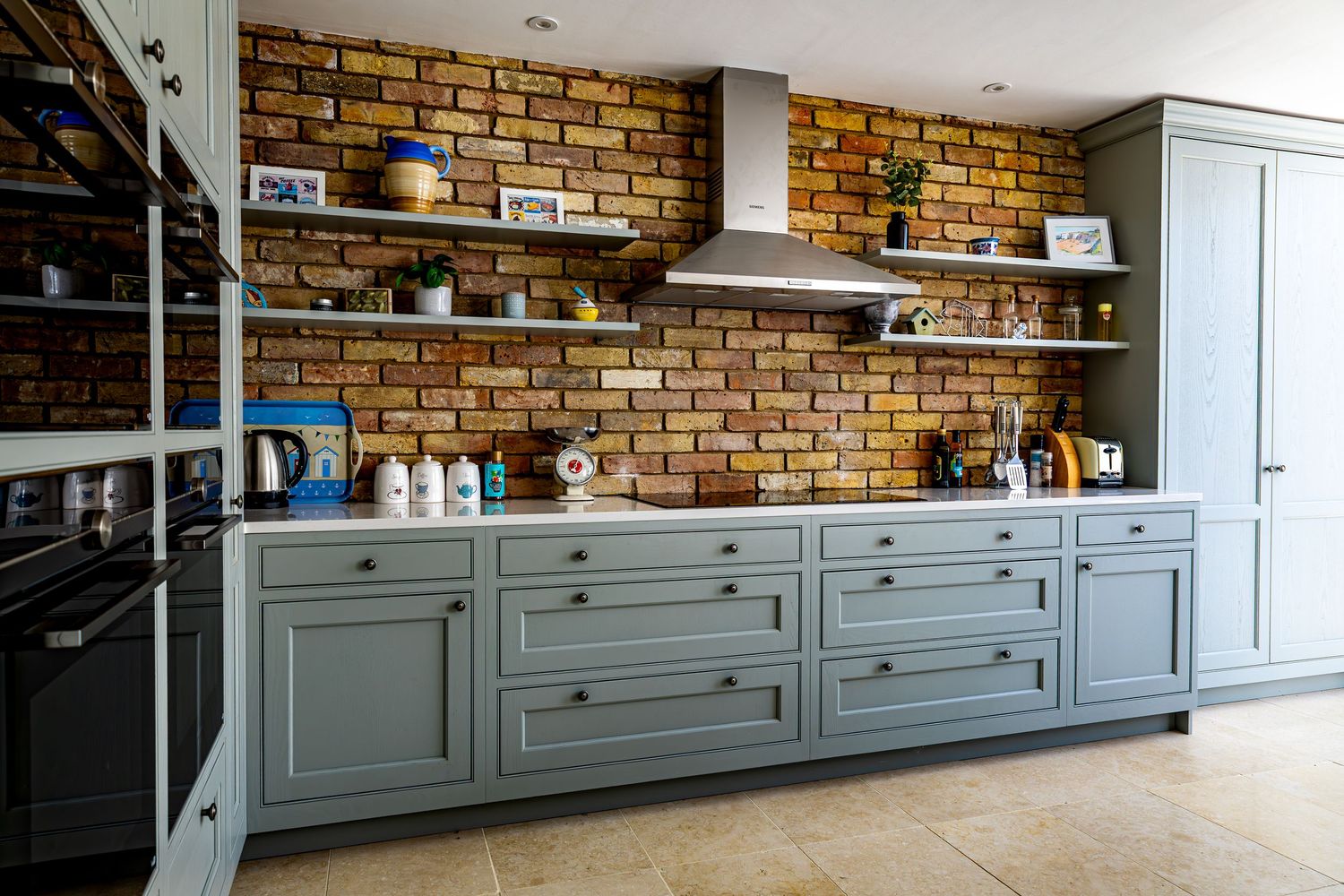 Stylish kitchen with sage green cabinets, exposed brick wall, open shelving, and range hood.