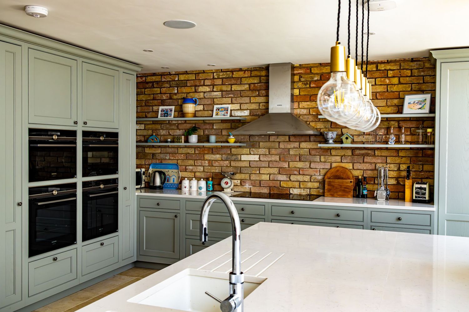 Contemporary kitchen with sage green cabinetry, white countertop island, exposed brick wall, and pendant lights.