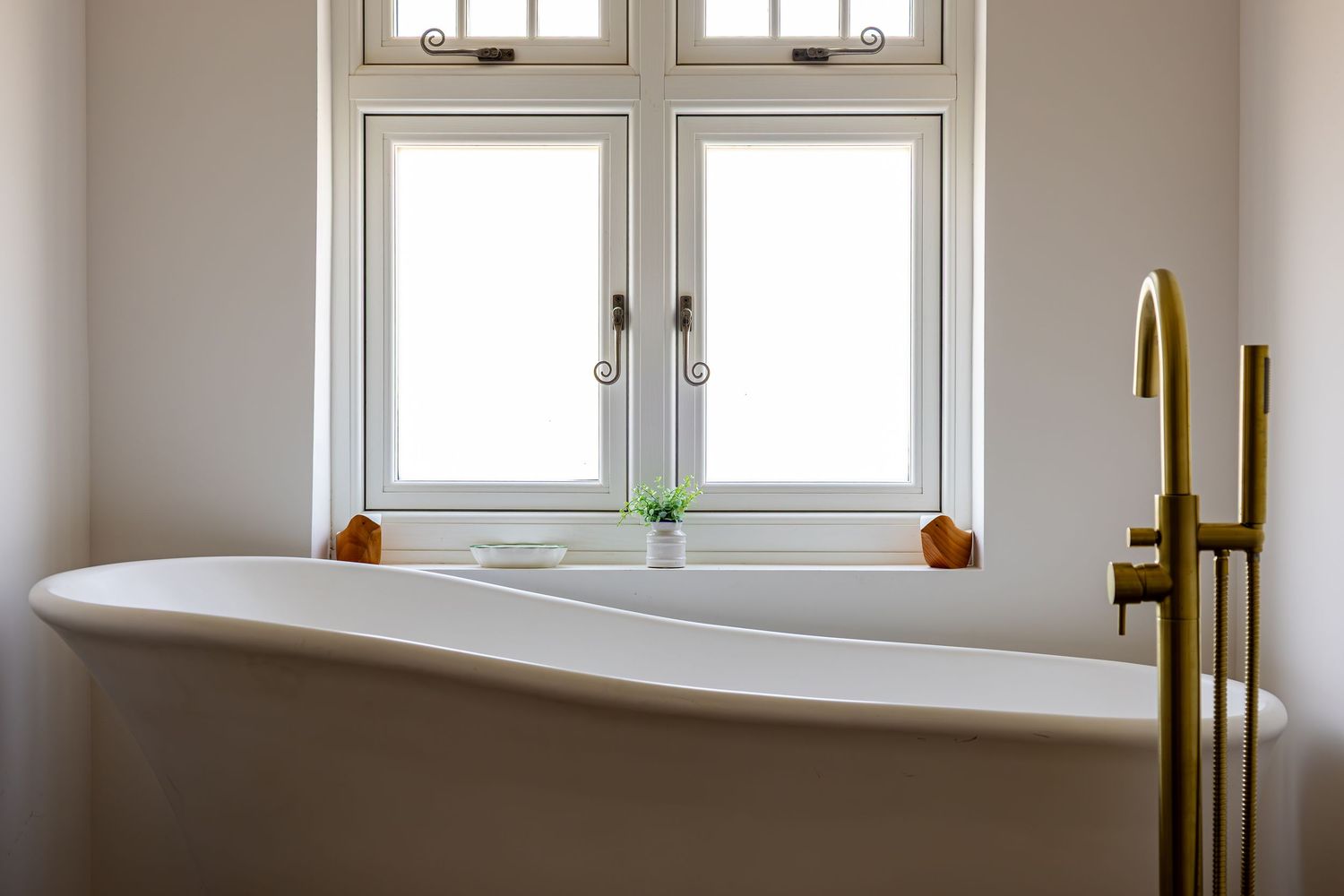 Elegant freestanding white bathtub with gold faucet beneath a bright double window in a minimalist bathroom.