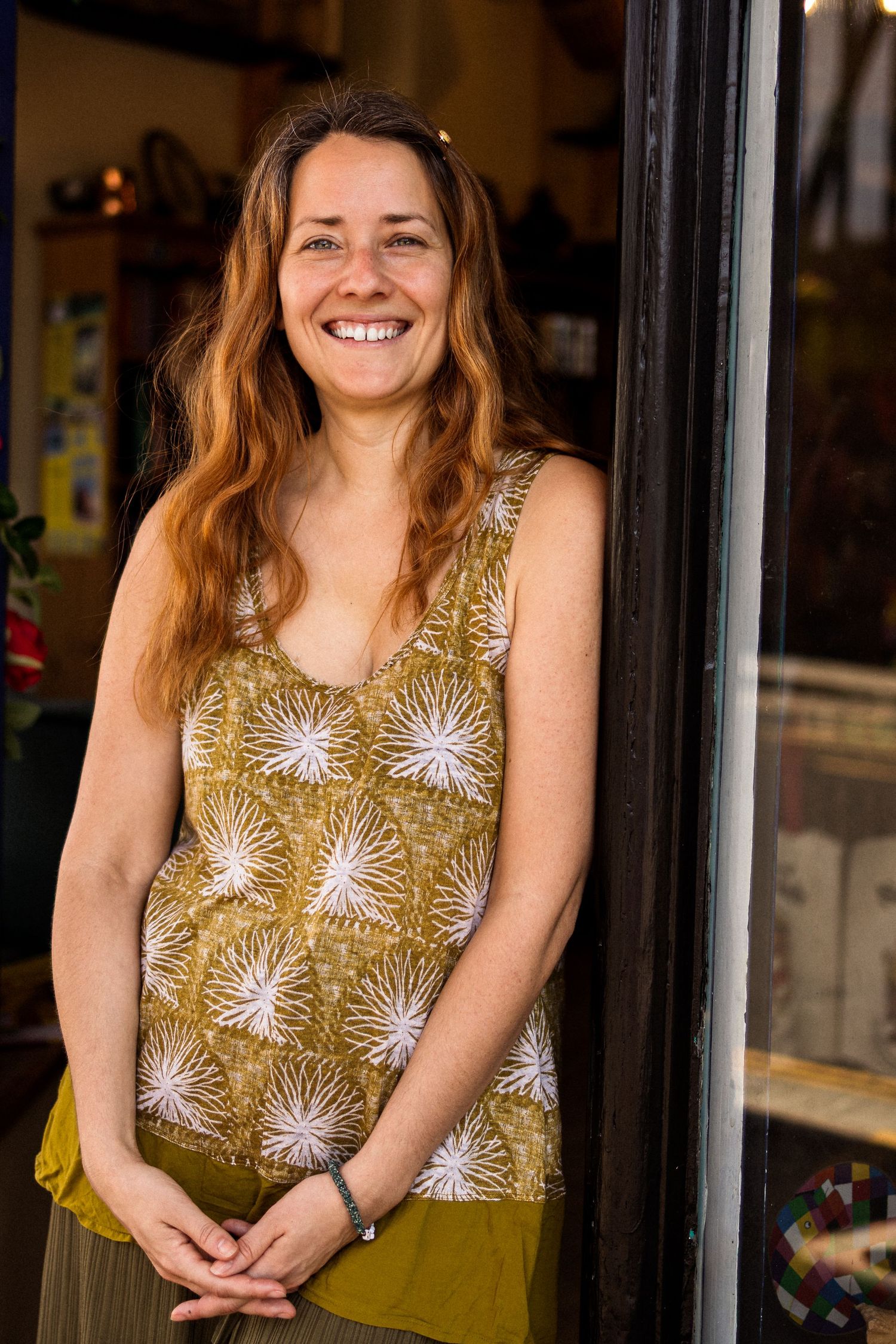 Smiling woman with auburn hair in olive floral sleeveless top leaning against doorframe of a shop.