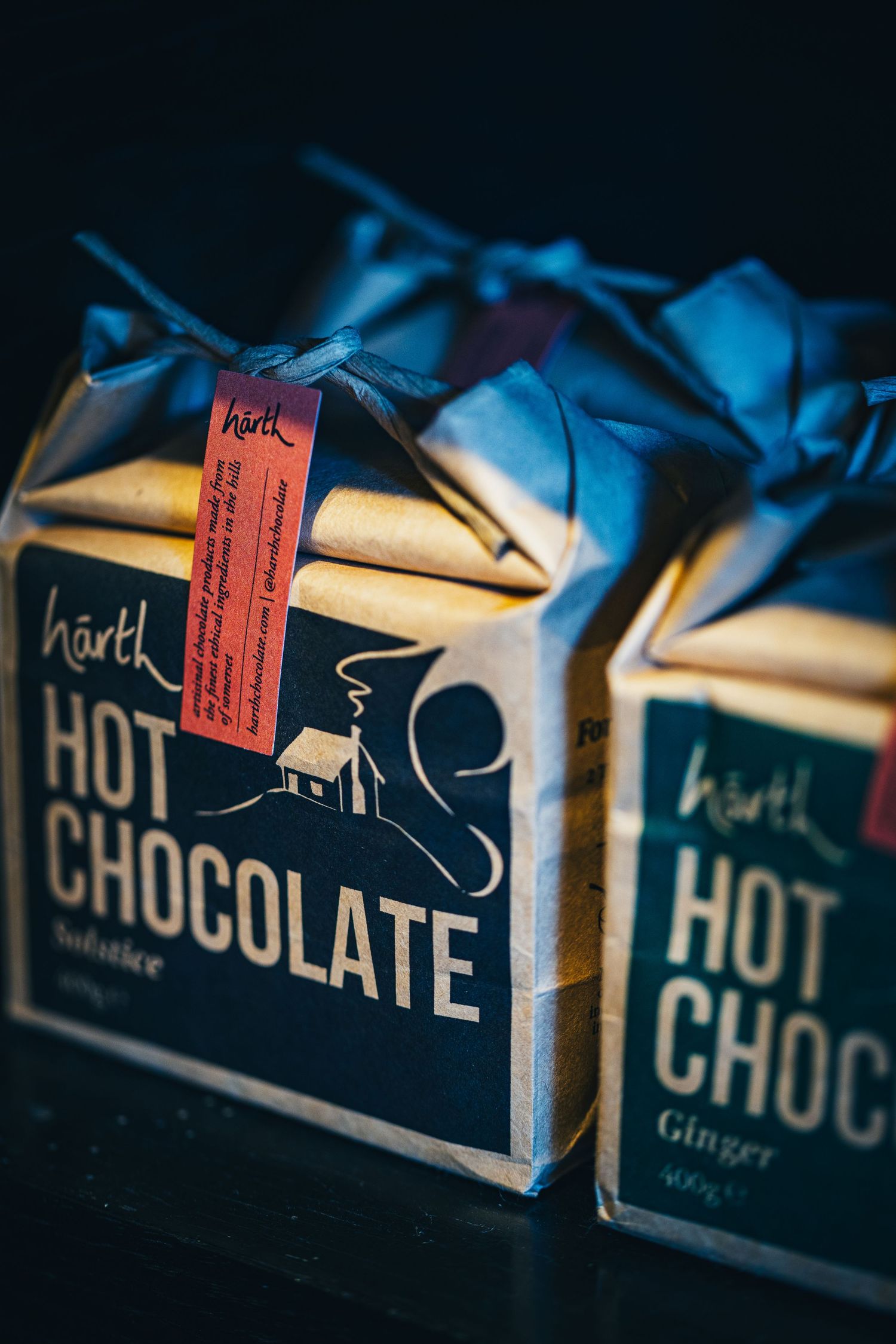Two Hartk Hot Chocolate packages in dark blue and brown kraft paper on a dark surface.