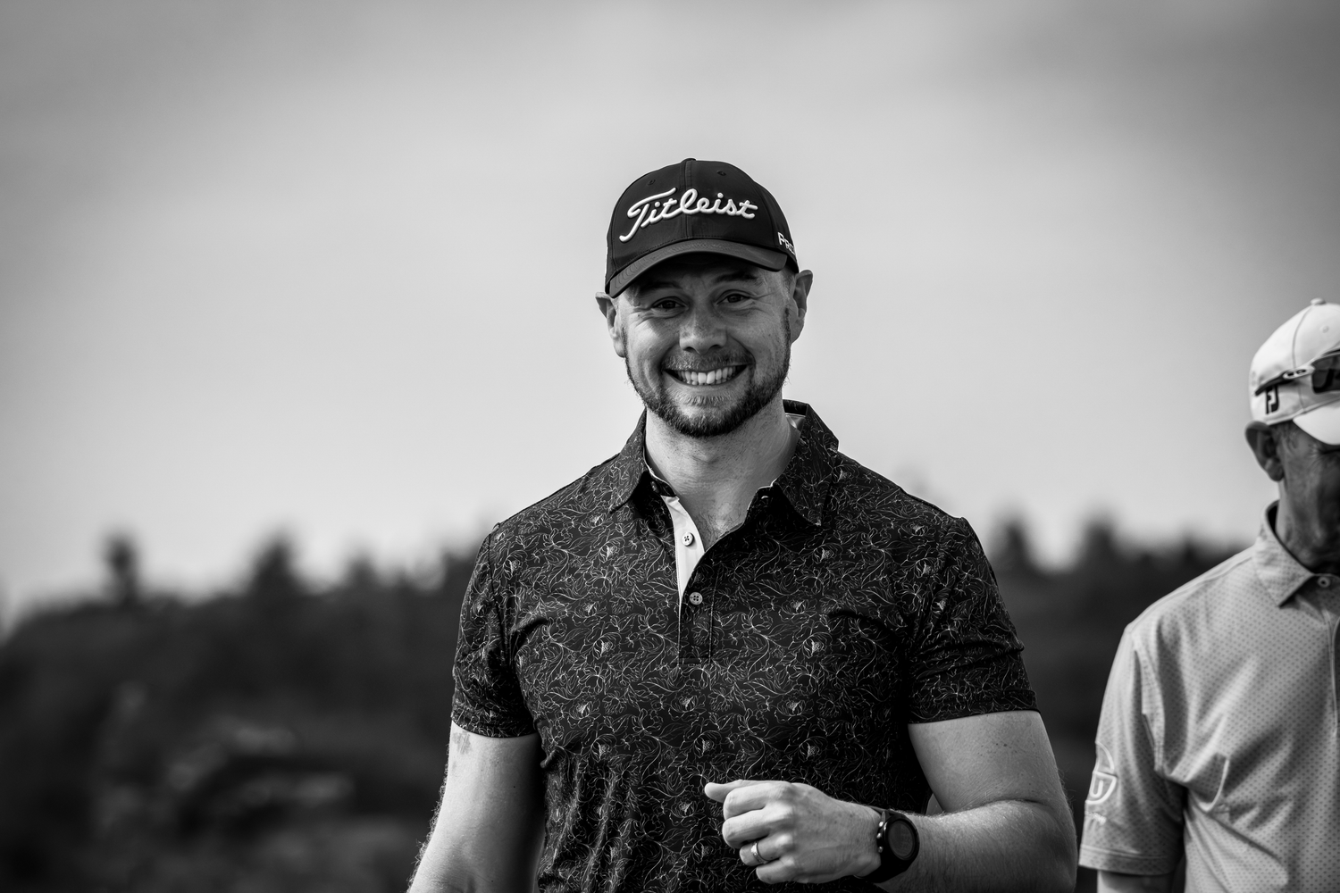 Happy man in Titleist cap and patterned polo shirt smiling outdoors in black and white photo.