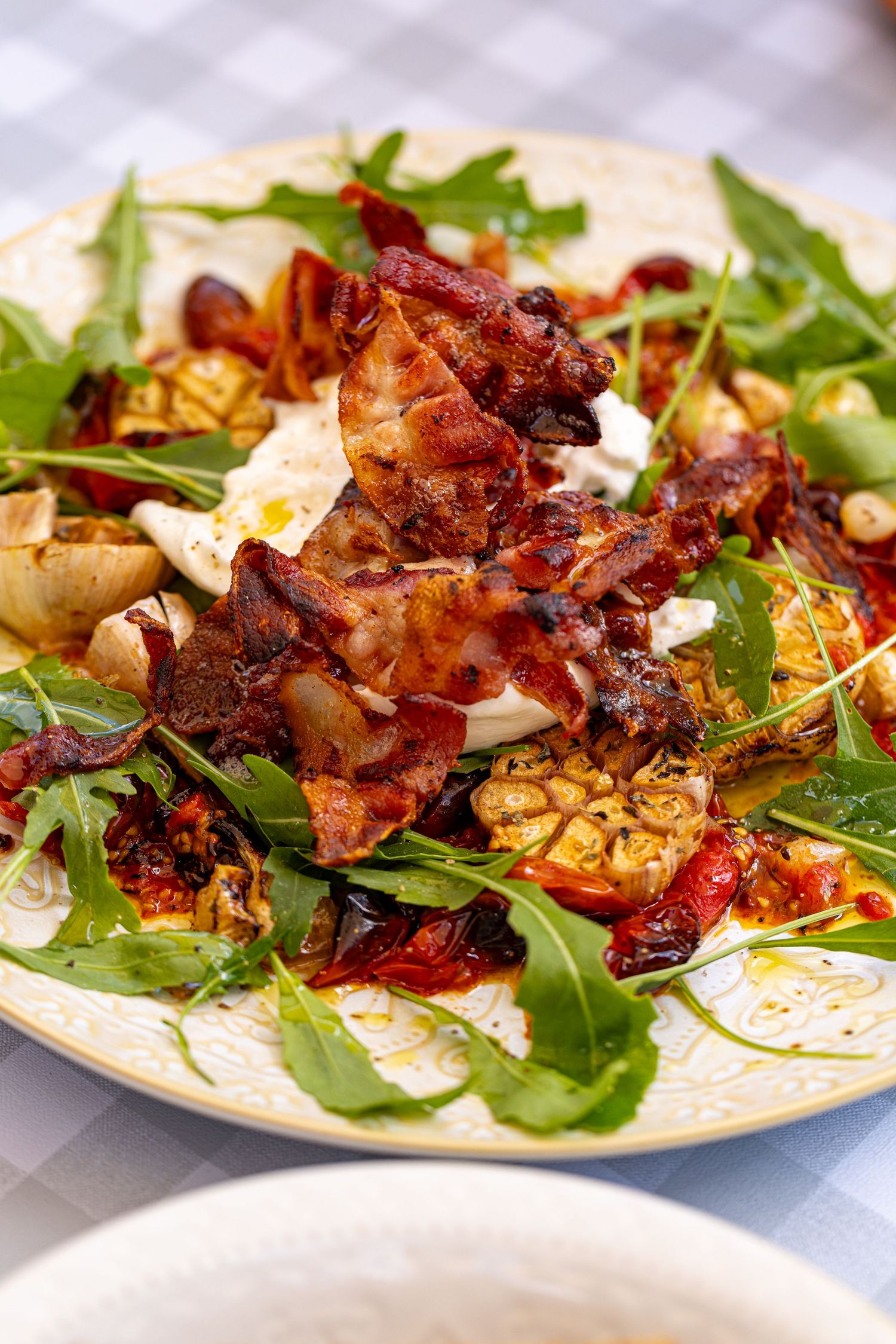Rustic salad plate with crispy bacon, burrata, roasted tomatoes, and fresh rocket on white tablecloth.