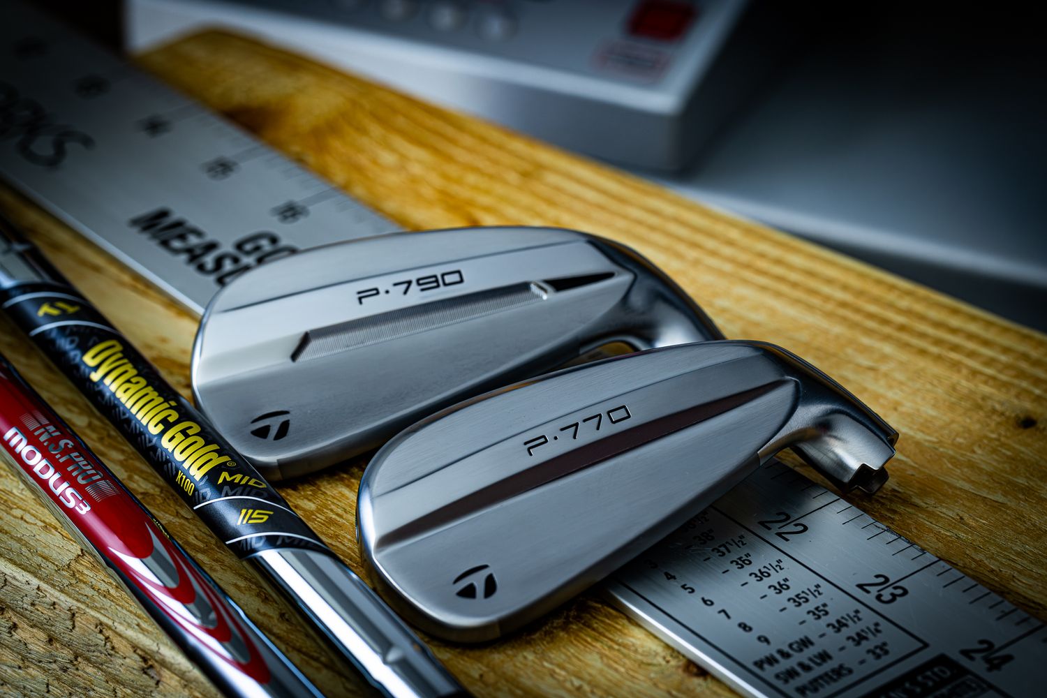 TaylorMade P-790 and P-770 golf irons displayed on a wooden surface with ruler and golf shafts.