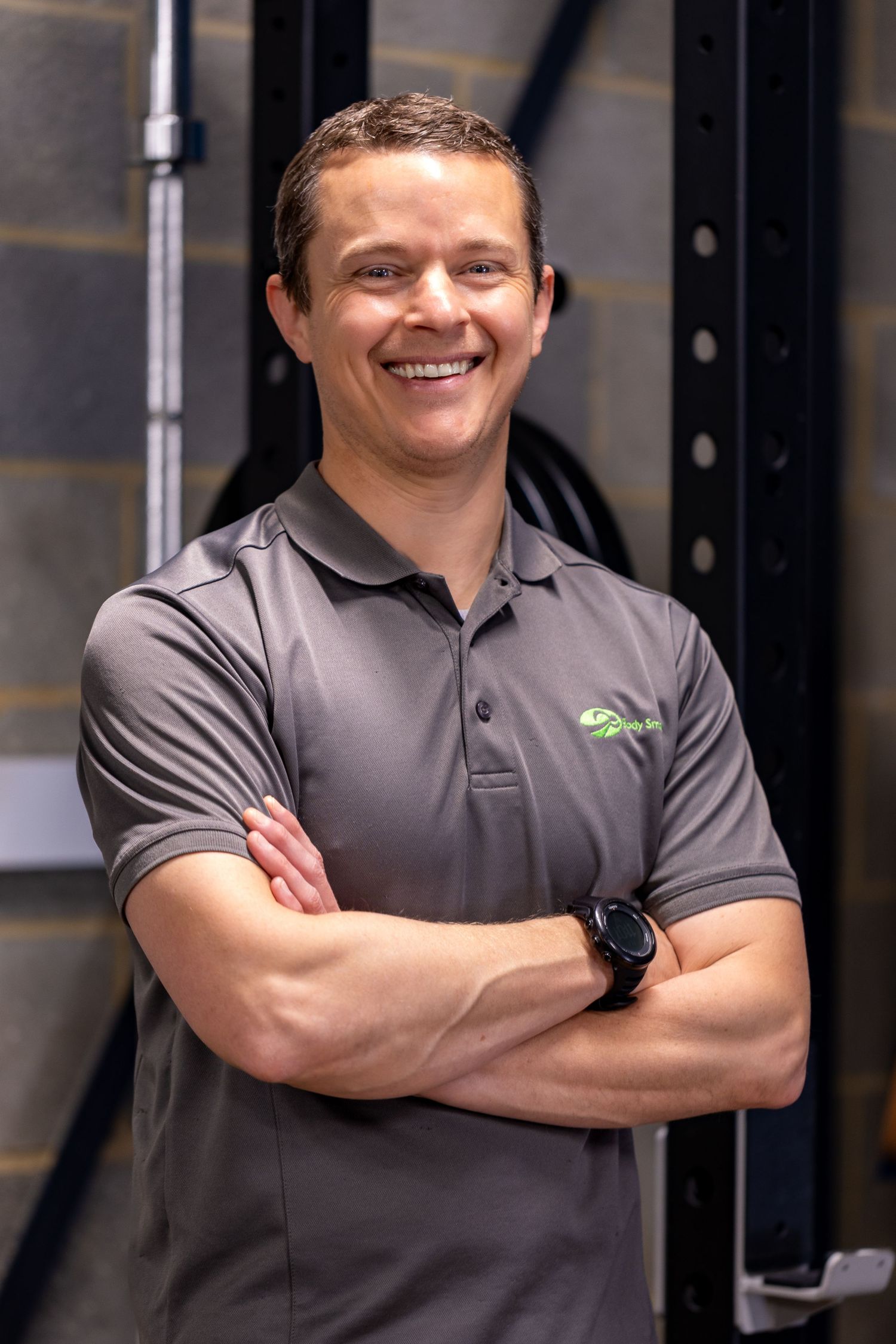 Smiling fitness trainer in gray polo shirt stands with arms crossed in a modern gym facility.
