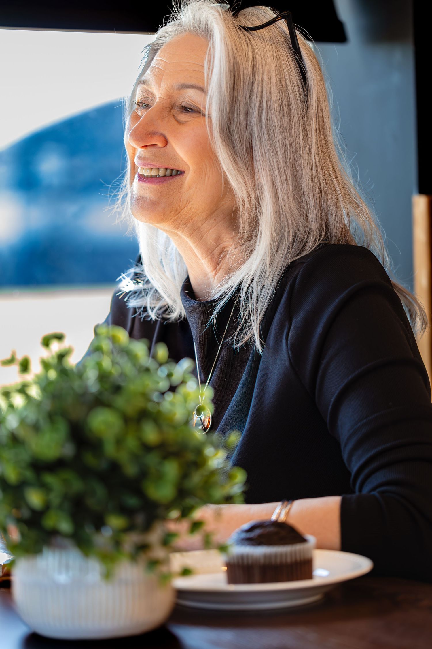 Smiling older woman with long white hair sitting at a cafe table with a chocolate dessert.