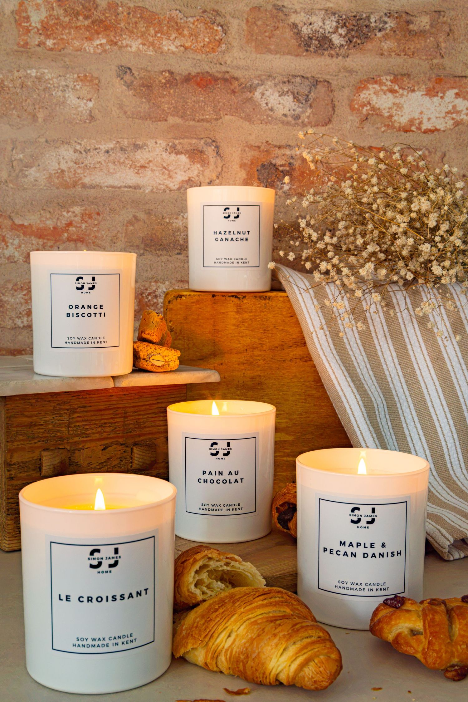 Four white soy wax candles with French pastry-inspired scents displayed with croissants on rustic brick background.