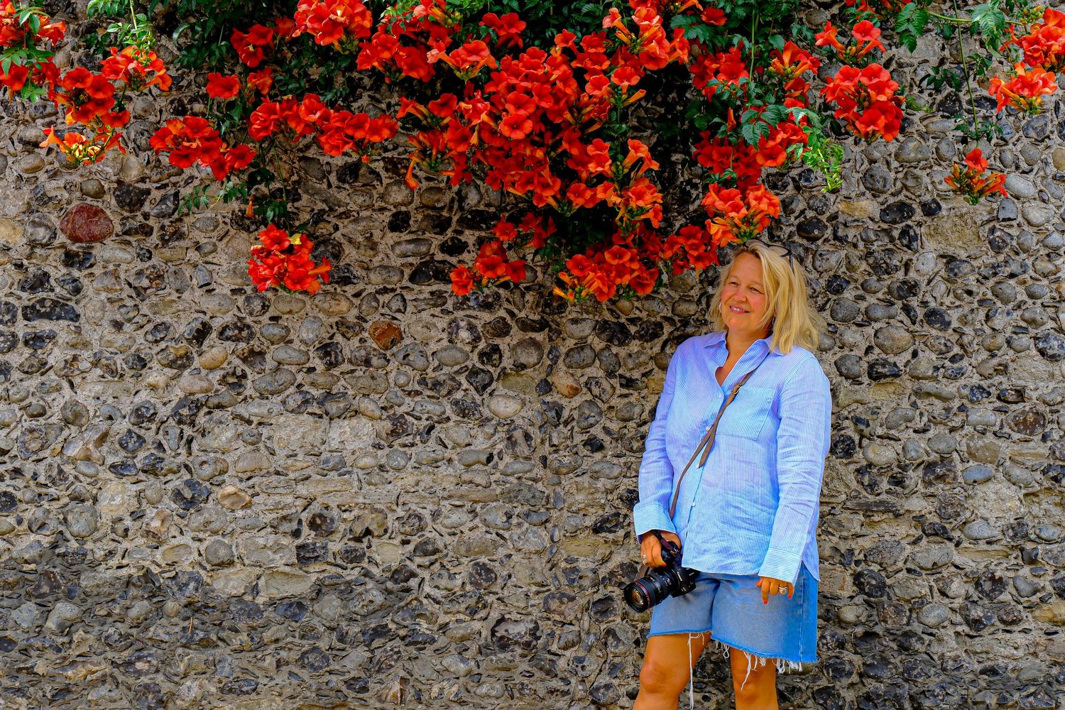 Woman in blue shirt standing against stone wall with vibrant red flowers overhead.