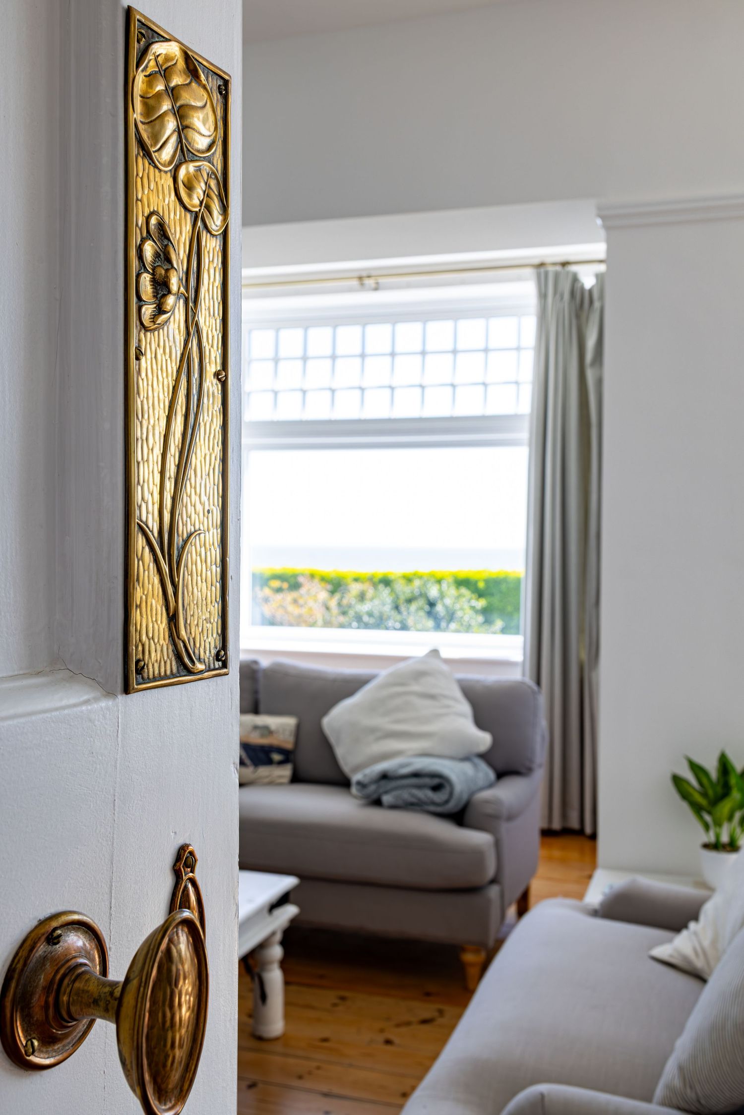 View through doorway with ornate brass handle into bright living room with grey sofa and window.