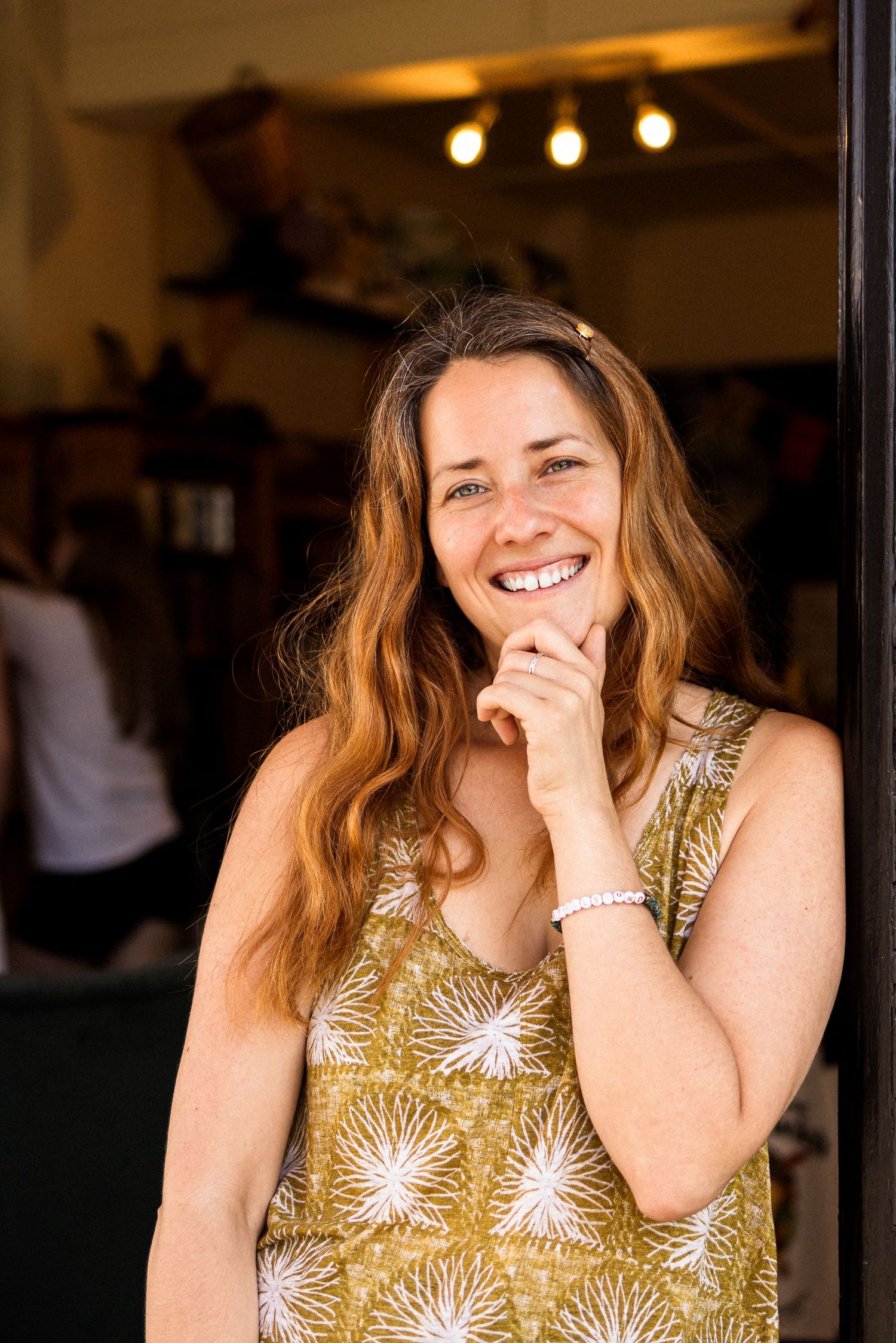 Smiling woman with auburn hair in floral dress standing in shop doorway.